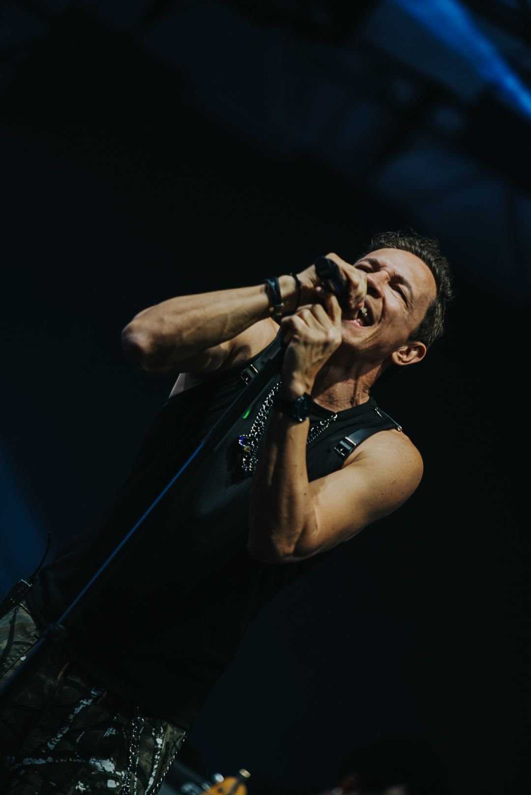 Um artista cantando ao microfone no palco; sorrindo com os braços erguidos, vestindo uma blusa sem mangas.