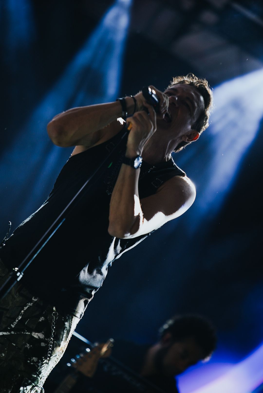 Cantor no palco, contraluz, segurando o microfone. Roupas escuras. Expressão intensa. Ambiente de concerto.