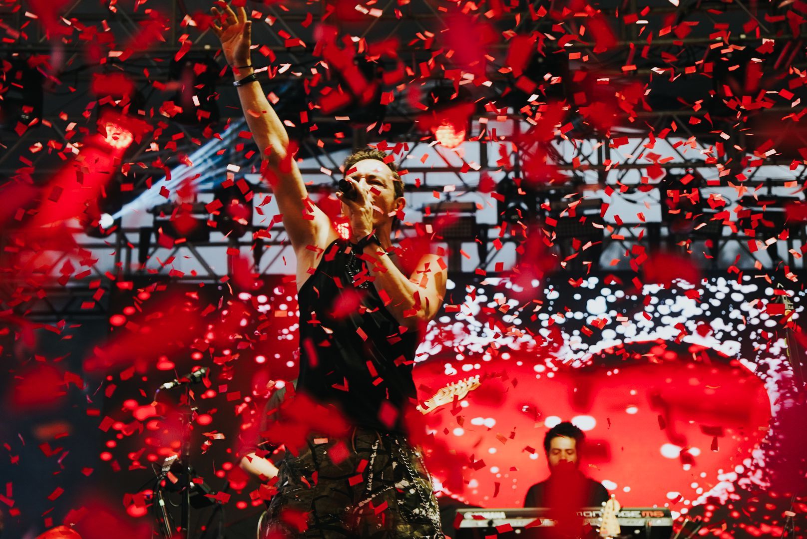 Cantor se apresentando no palco, braços erguidos, confetes caindo; fundo vermelho com um coração.