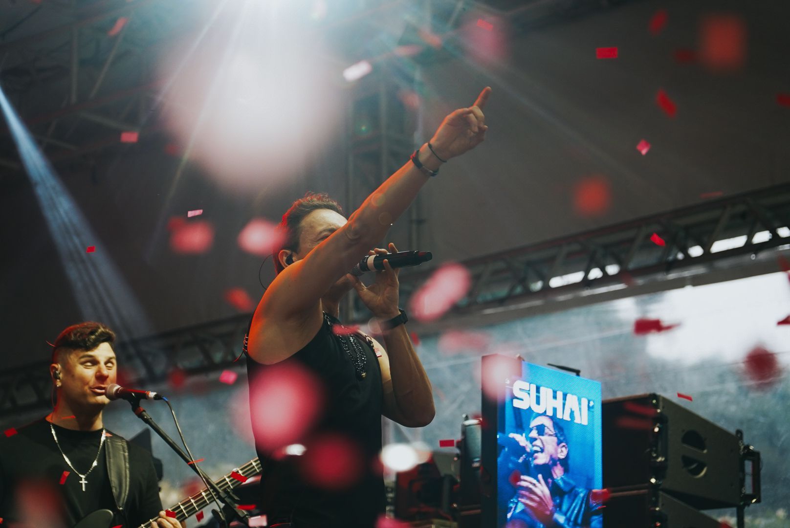 Um artista cantando no palco, braço erguido, confetes caindo. Outro músico toca violão. Cenário de palco.