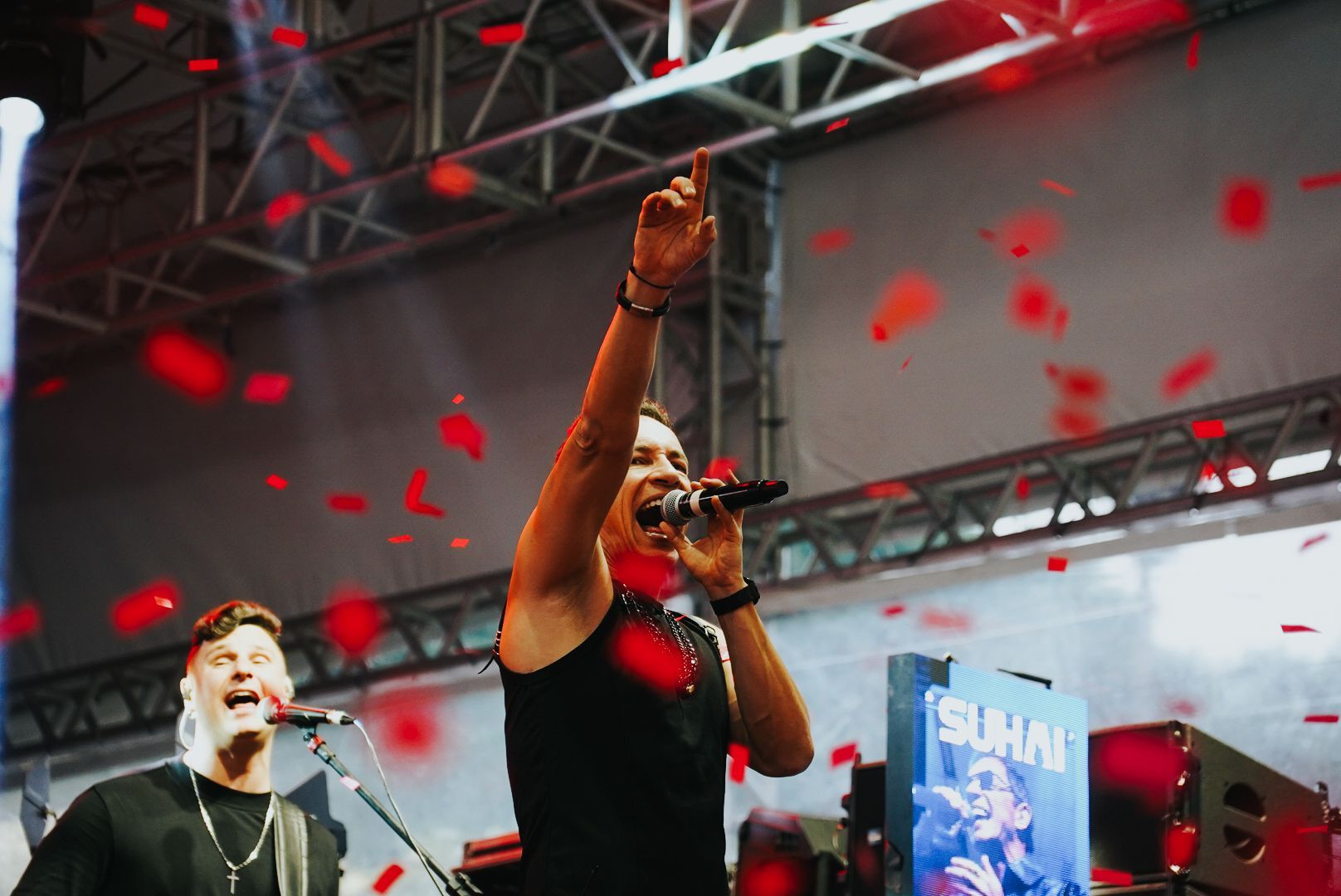 Um artista no palco com o braço levantado, confetes caindo. Outro músico ao lado dele canta em um microfone.