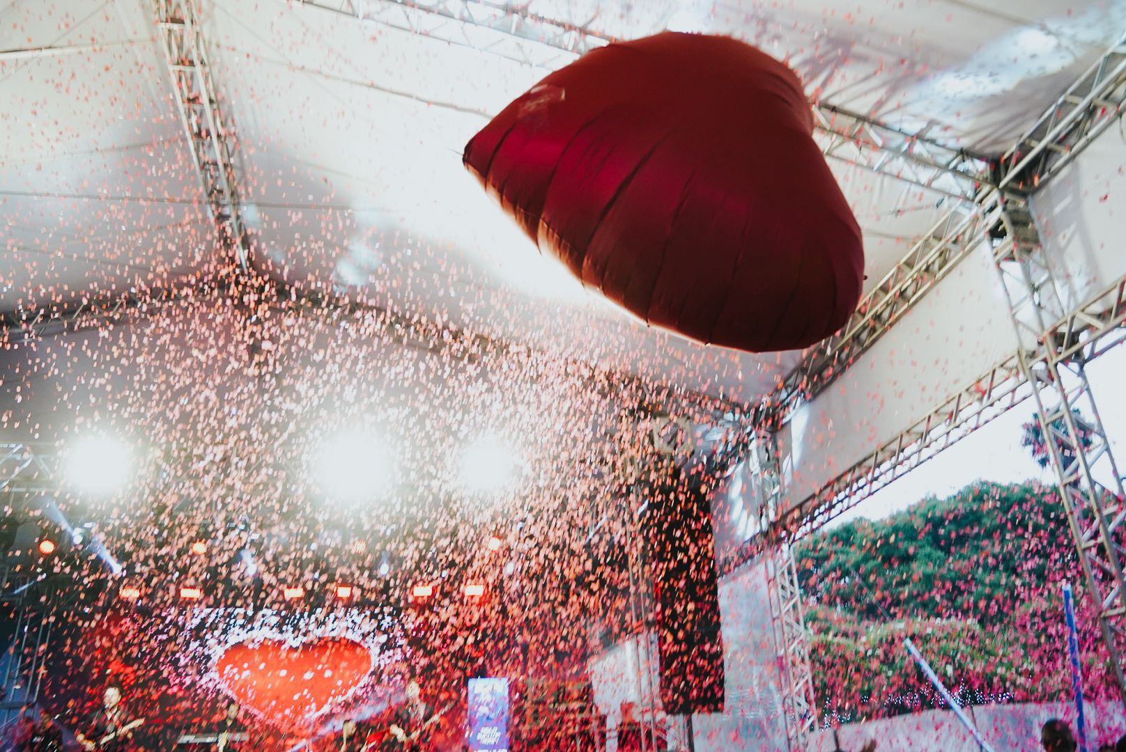 Balão vermelho em forma de coração subindo com confetes em um evento ao ar livre, com palco e luzes.