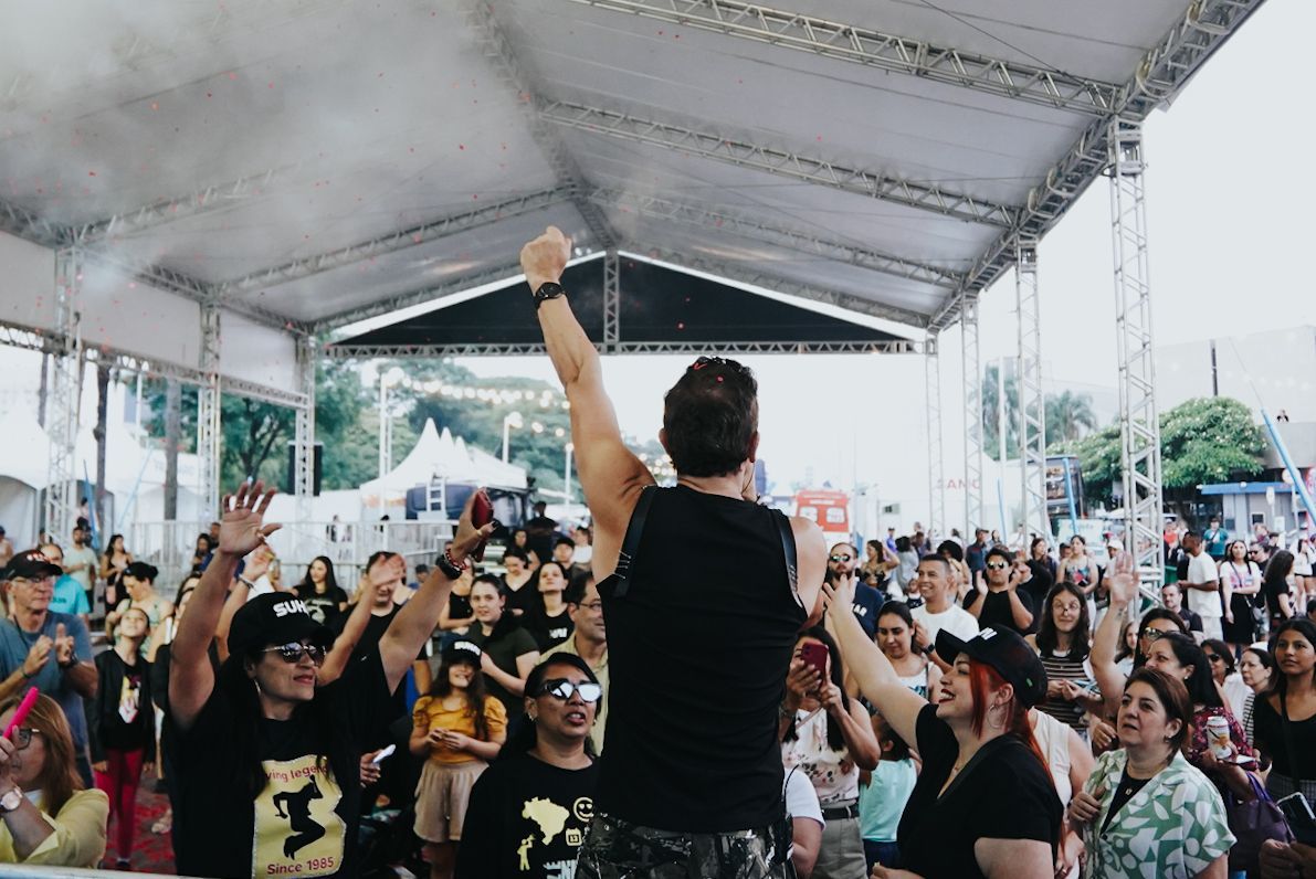Músico no palco com o braço levantado, apresentando-se para a plateia em evento ao ar livre sob uma tenda.