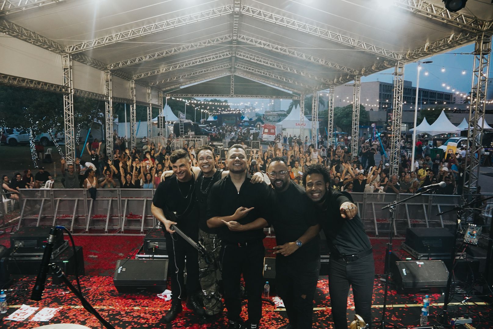 A banda está no palco, de braços dados, de frente para a plateia em um ambiente ao ar livre.