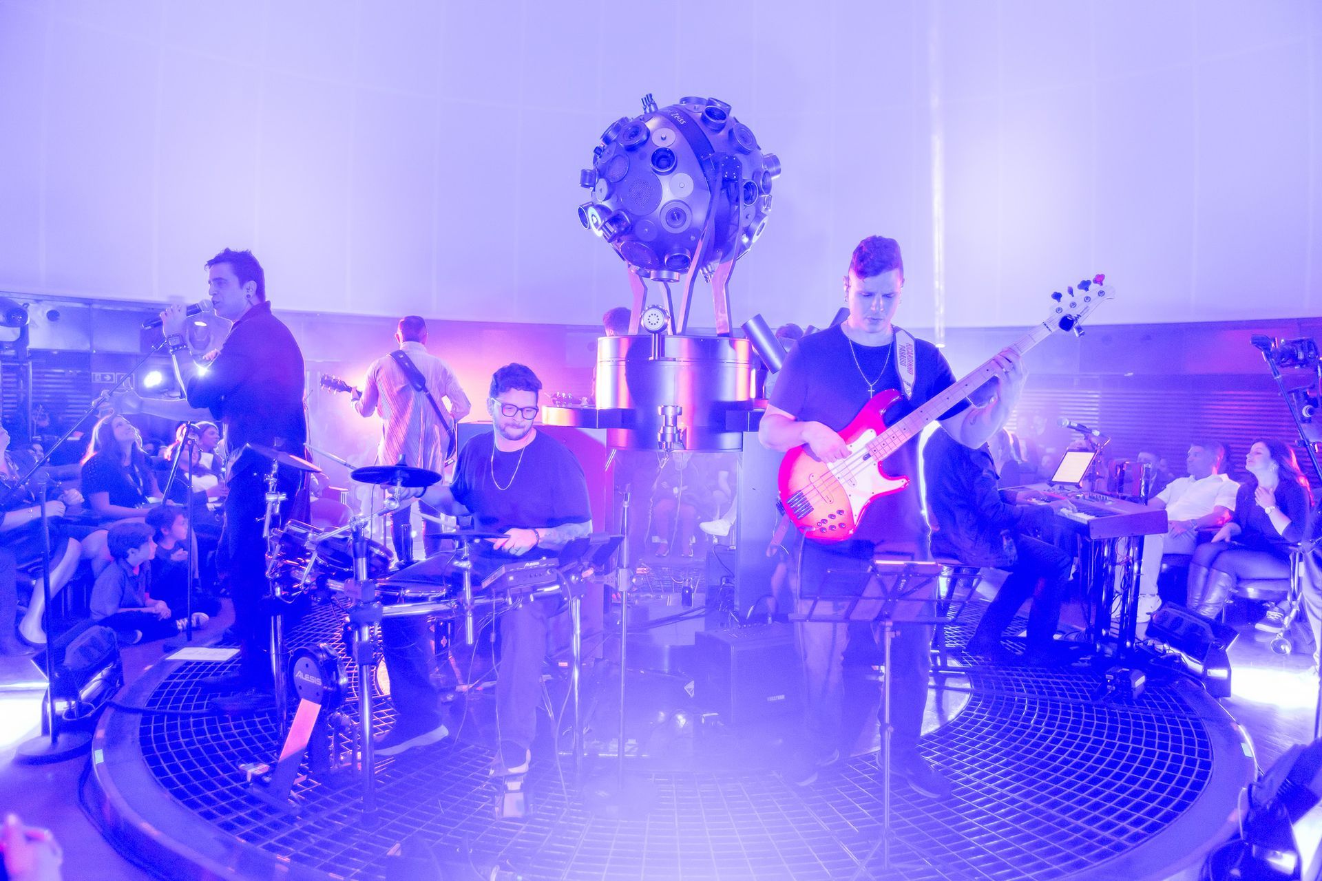 Uma banda toca instrumentos em um palco circular banhado por uma luz roxa brilhante, com uma grande estrutura esférica 