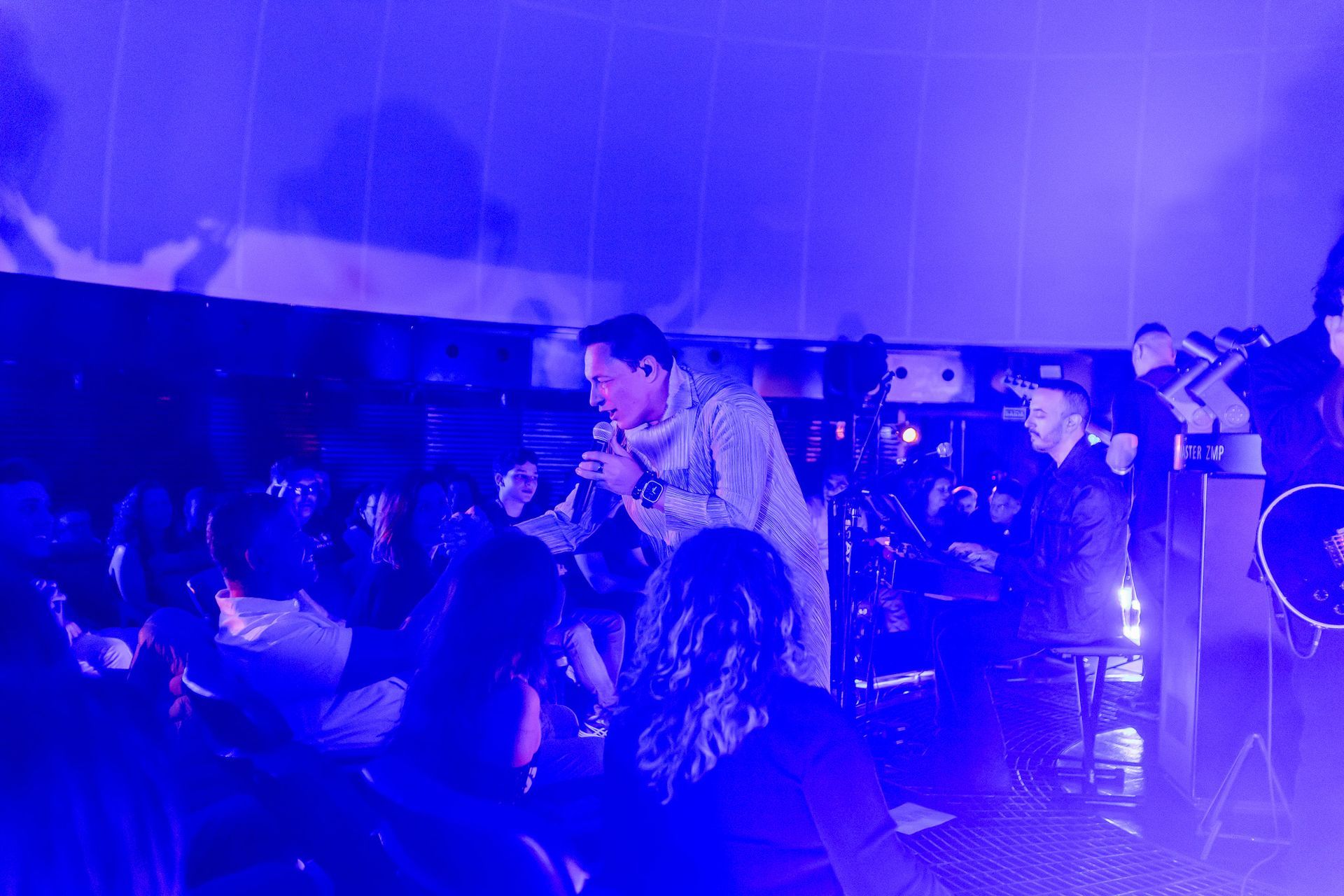 Um artista canta em um microfone no palco, banhado por luz azul, enquanto o público assiste na escuridão.