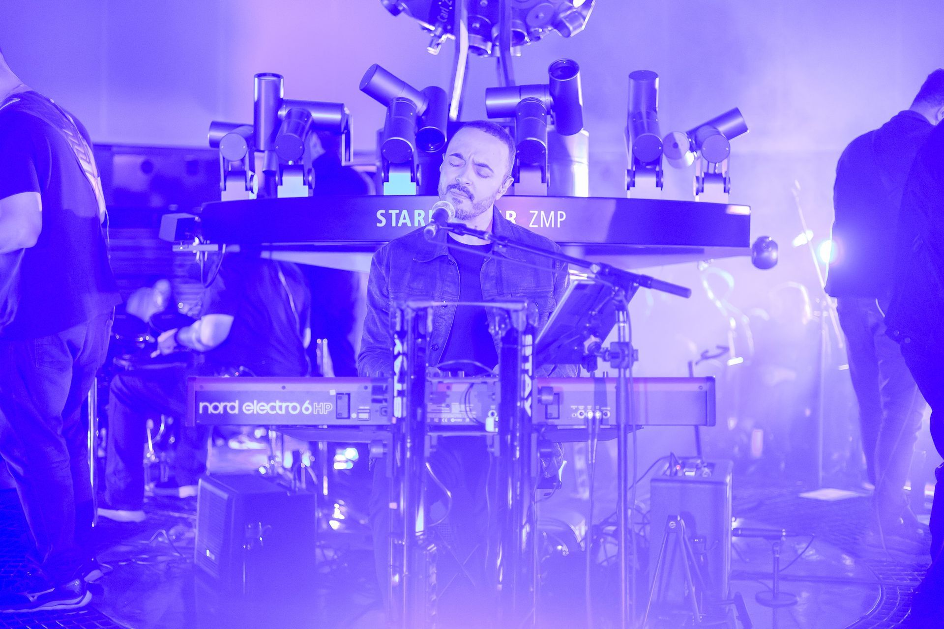 Um tecladista se apresenta no palco, iluminado por uma luz azul brilhante em meio a uma estrutura de show.