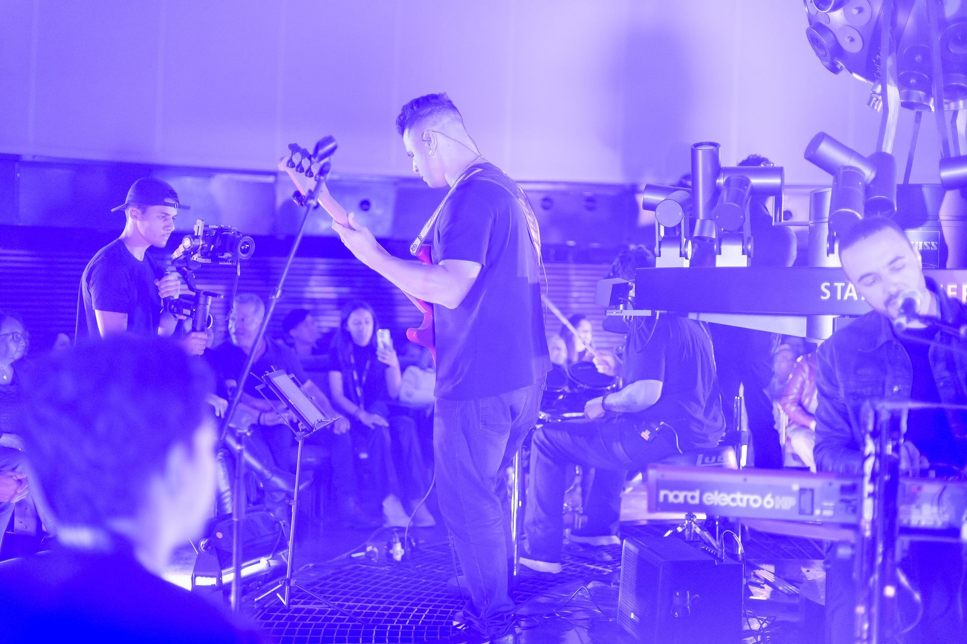 Uma banda se apresenta no palco sob iluminação roxa, com um guitarrista no centro e um tecladista à direita.