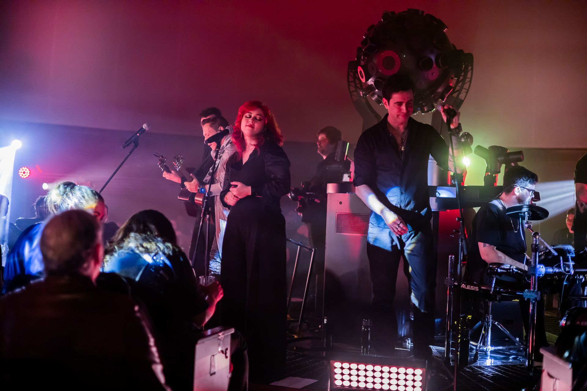 Uma banda se apresenta em um palco, composta por um vocalista, um guitarrista, um tecladista e um baterista.