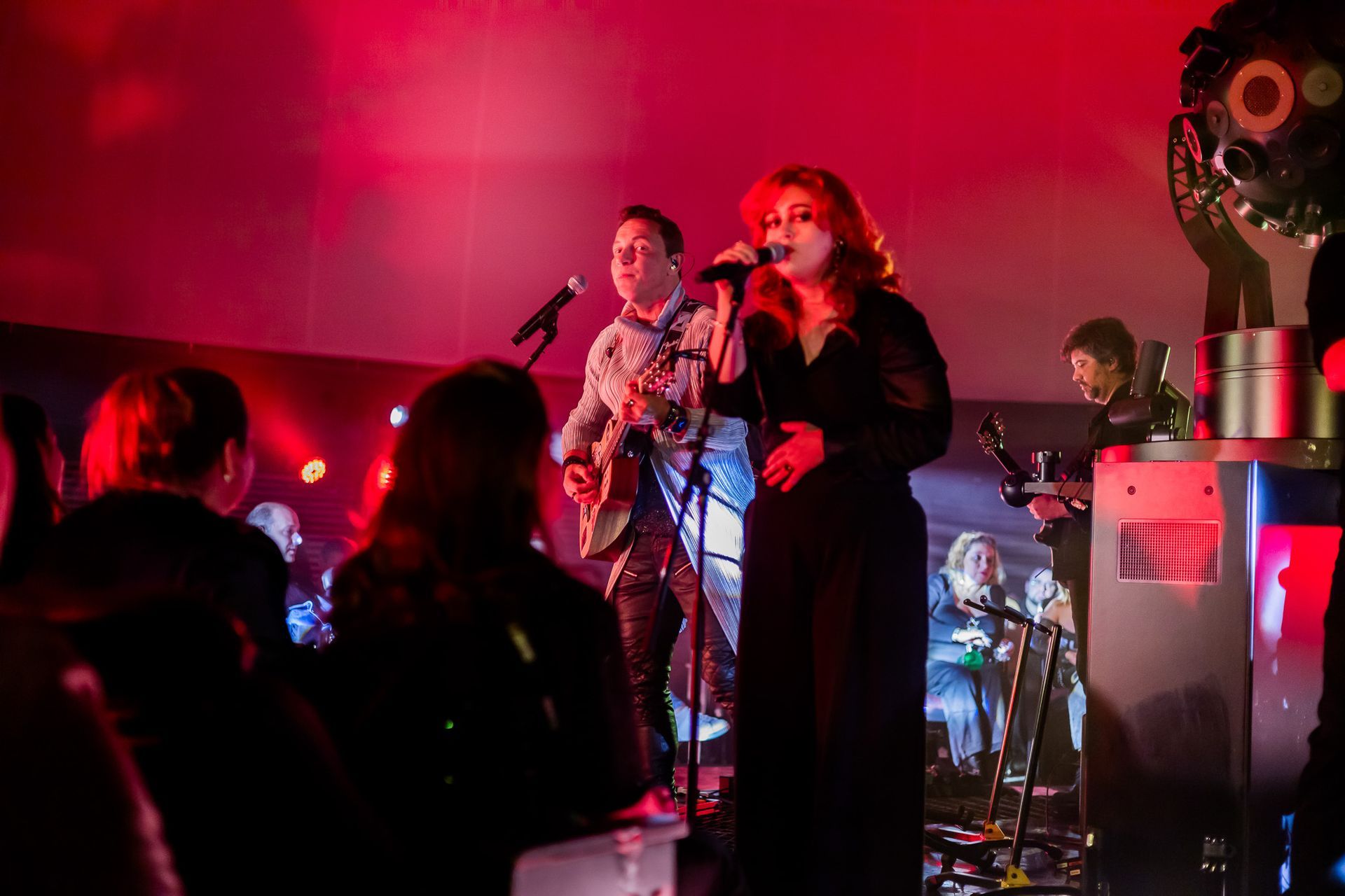 Uma banda se apresenta no palco sob iluminação vermelha, com um vocalista segurando um microfone e um músico tocando violão.