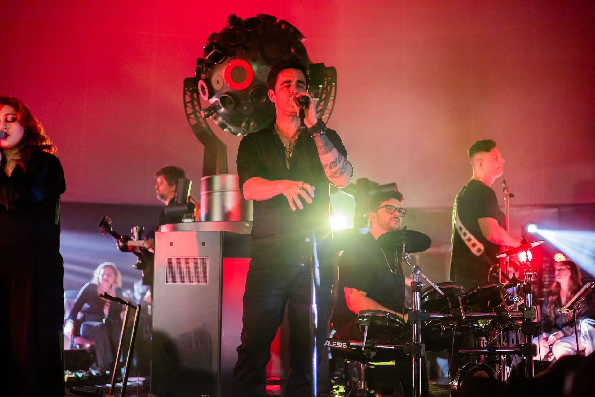 Uma banda se apresenta no palco sob iluminação vermelha, com o vocalista principal no centro do palco cantando.