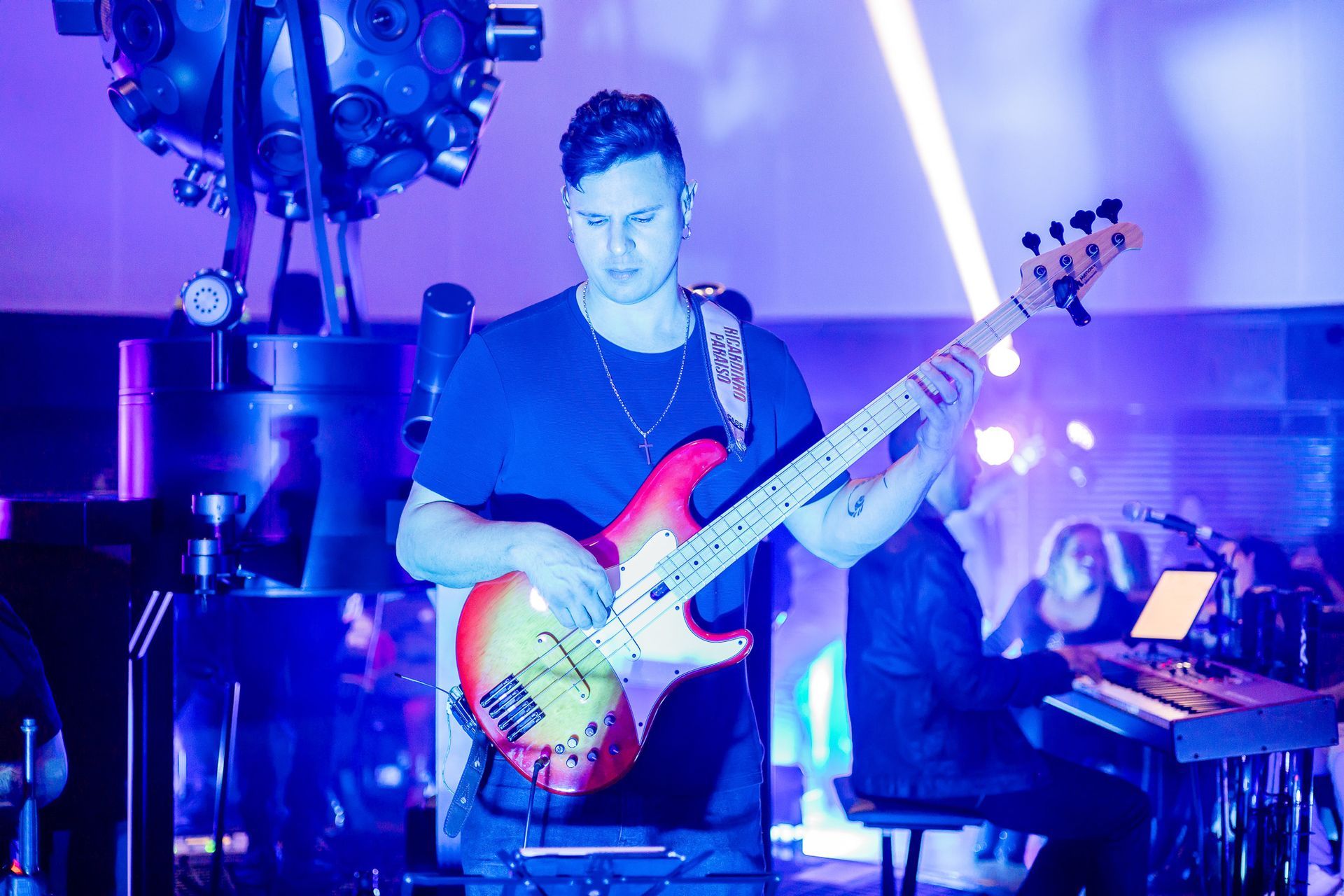 Um baixista se apresenta no palco sob luz azul ao lado de um tecladista, com um grande sistema de iluminação ao fundo.