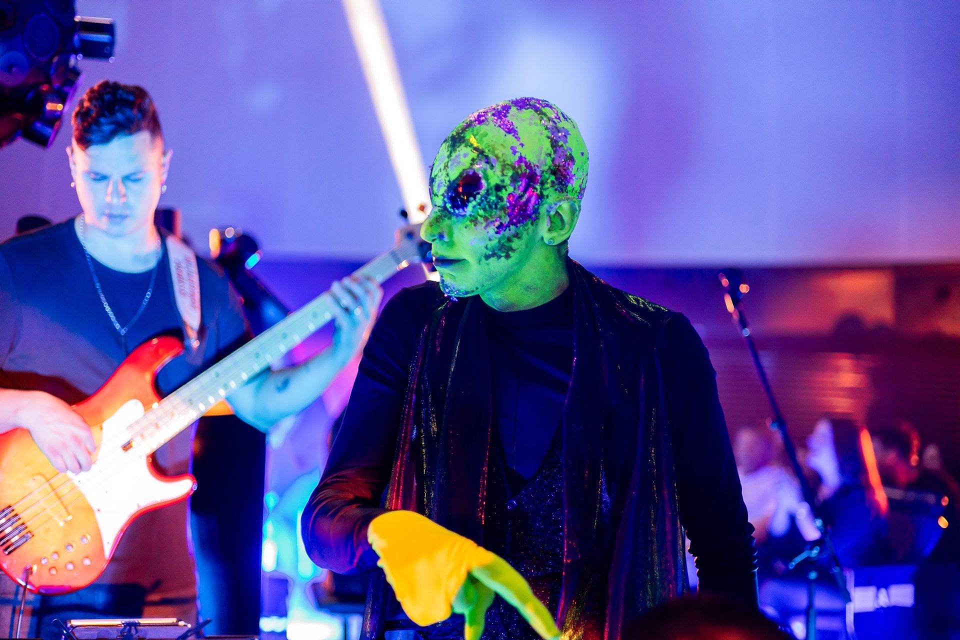 Um artista com o rosto e a cabeça pintados de verde está ao lado de um baixista em um palco pouco iluminado com luzes roxas.