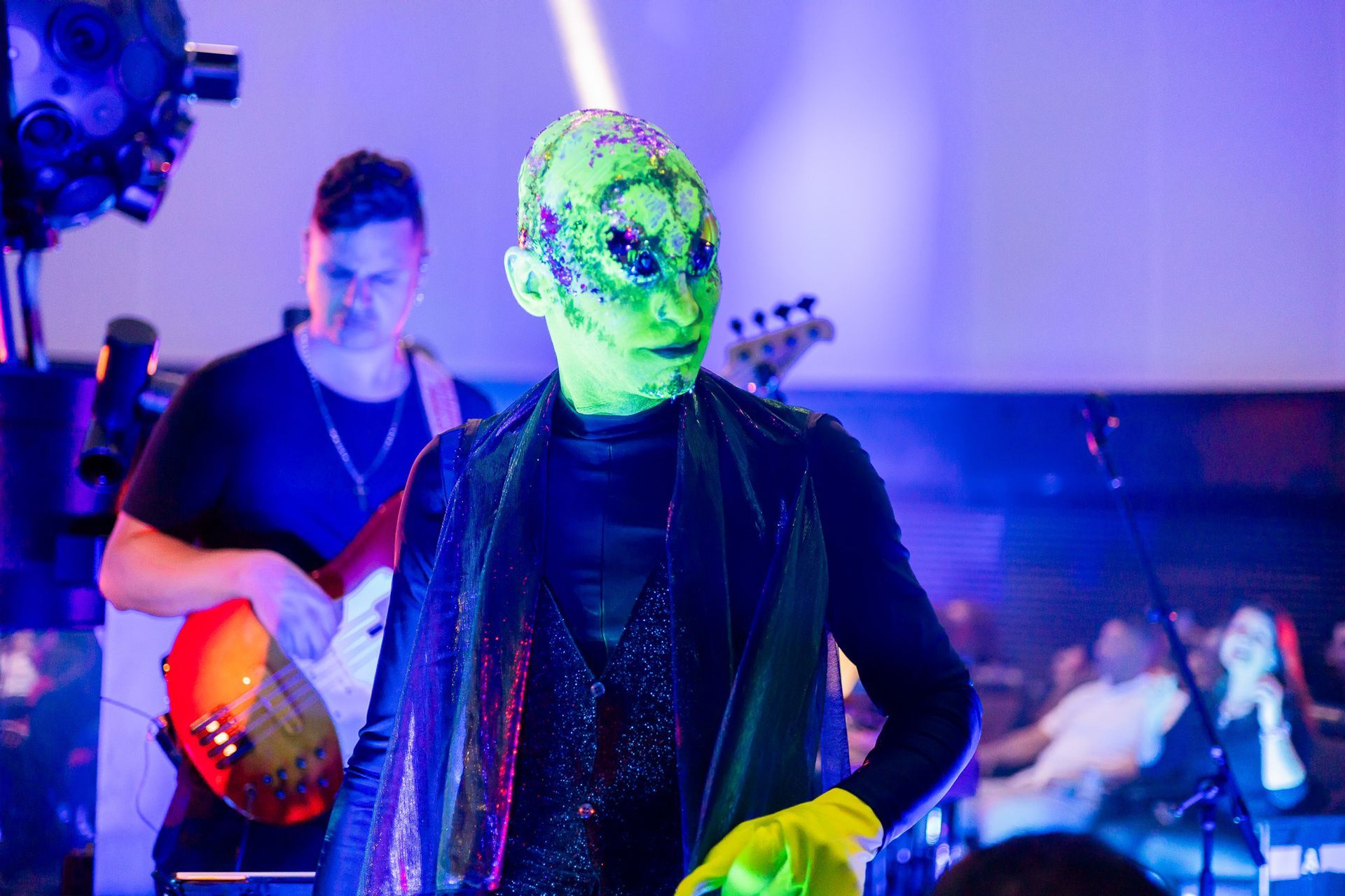 Um artista usando uma máscara texturizada verde neon e roupas escuras está no palco em frente a um baixista durante um show.