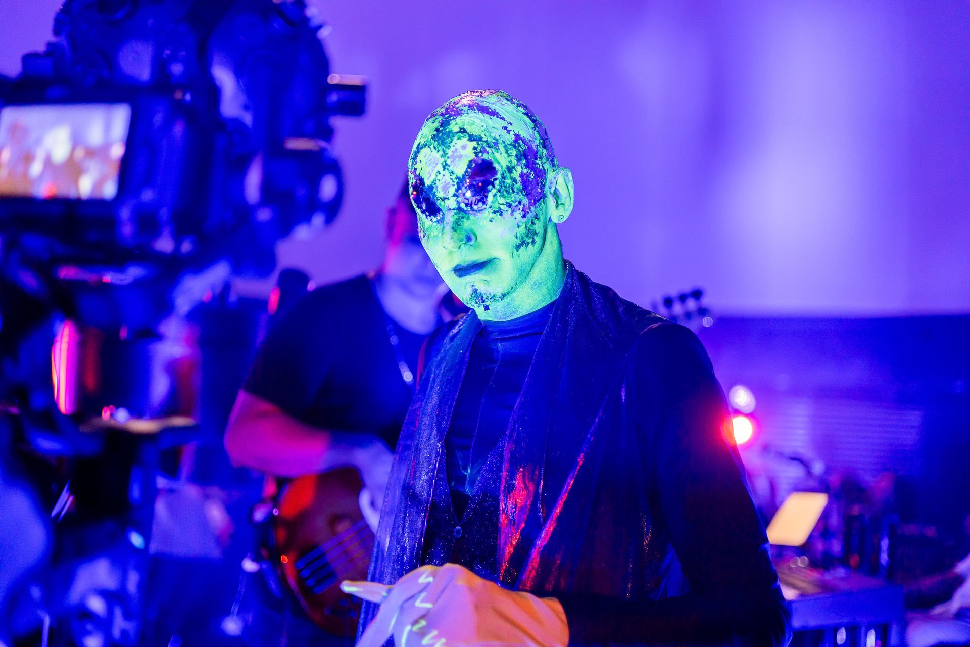 Um artista com maquiagem facial elaborada e fluorescente se apresenta em um local pouco iluminado, perto de um músico.