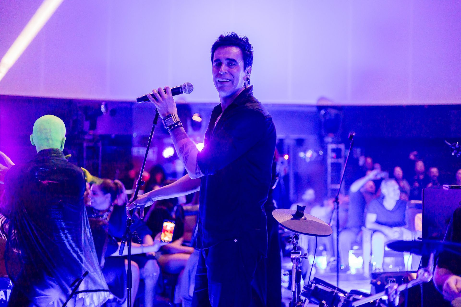 Um artista canta em um microfone em um palco iluminado com tons roxos brilhantes, com uma plateia visível ao fundo.