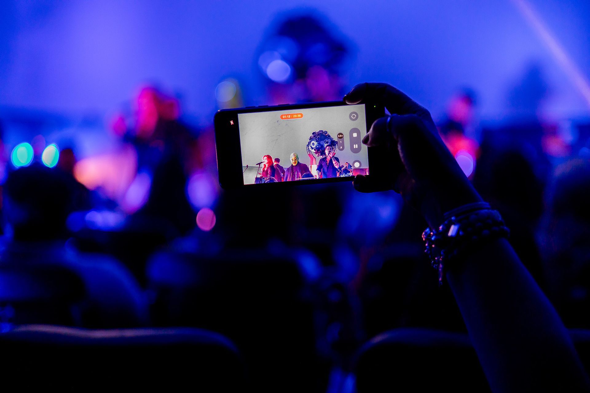 Uma pessoa segura um celular em um local escuro com iluminação azul, filmando uma apresentação no palco.