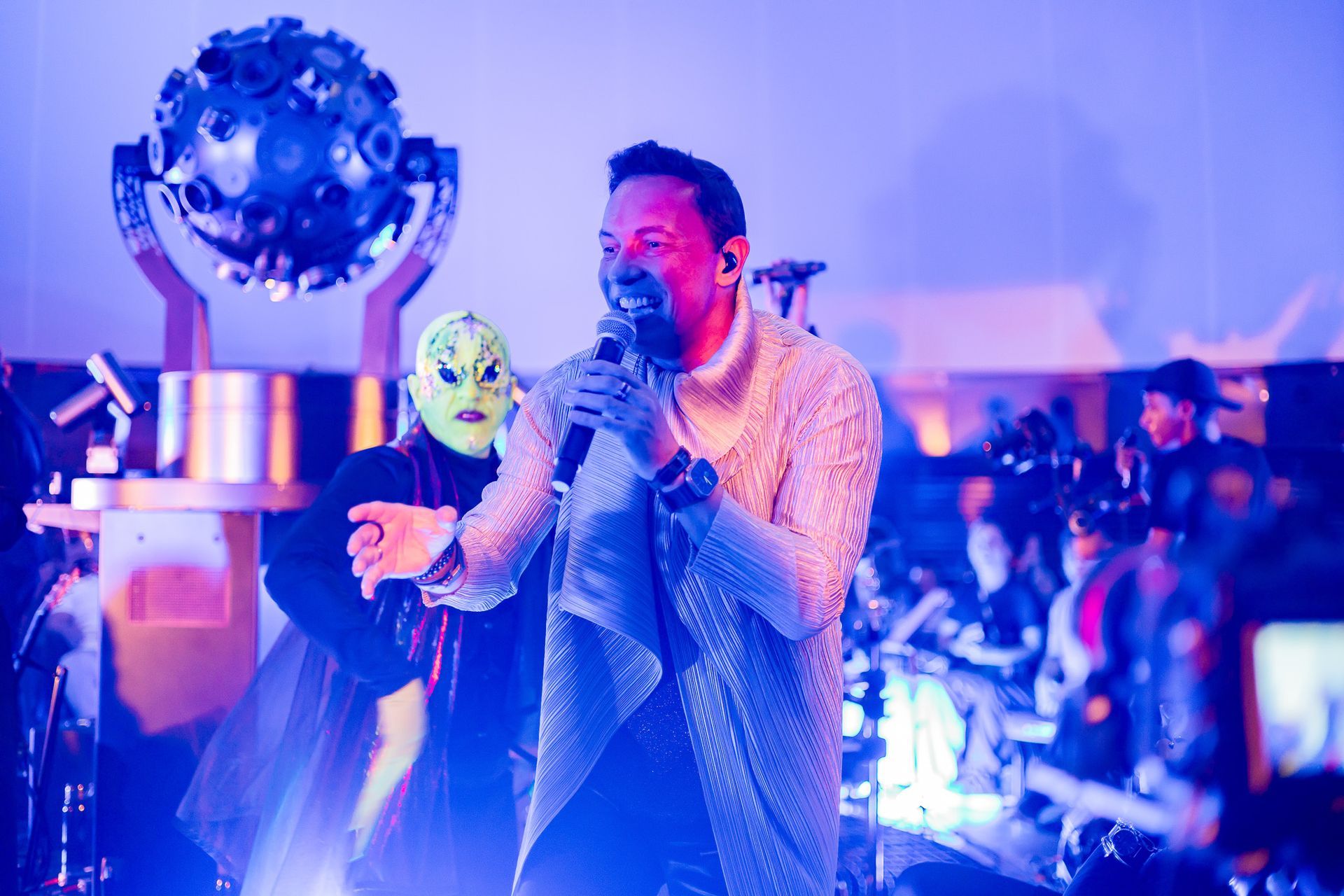 Um artista com uma jaqueta texturizada canta em um microfone no palco, ao lado de um artista usando uma máscara estilizada.