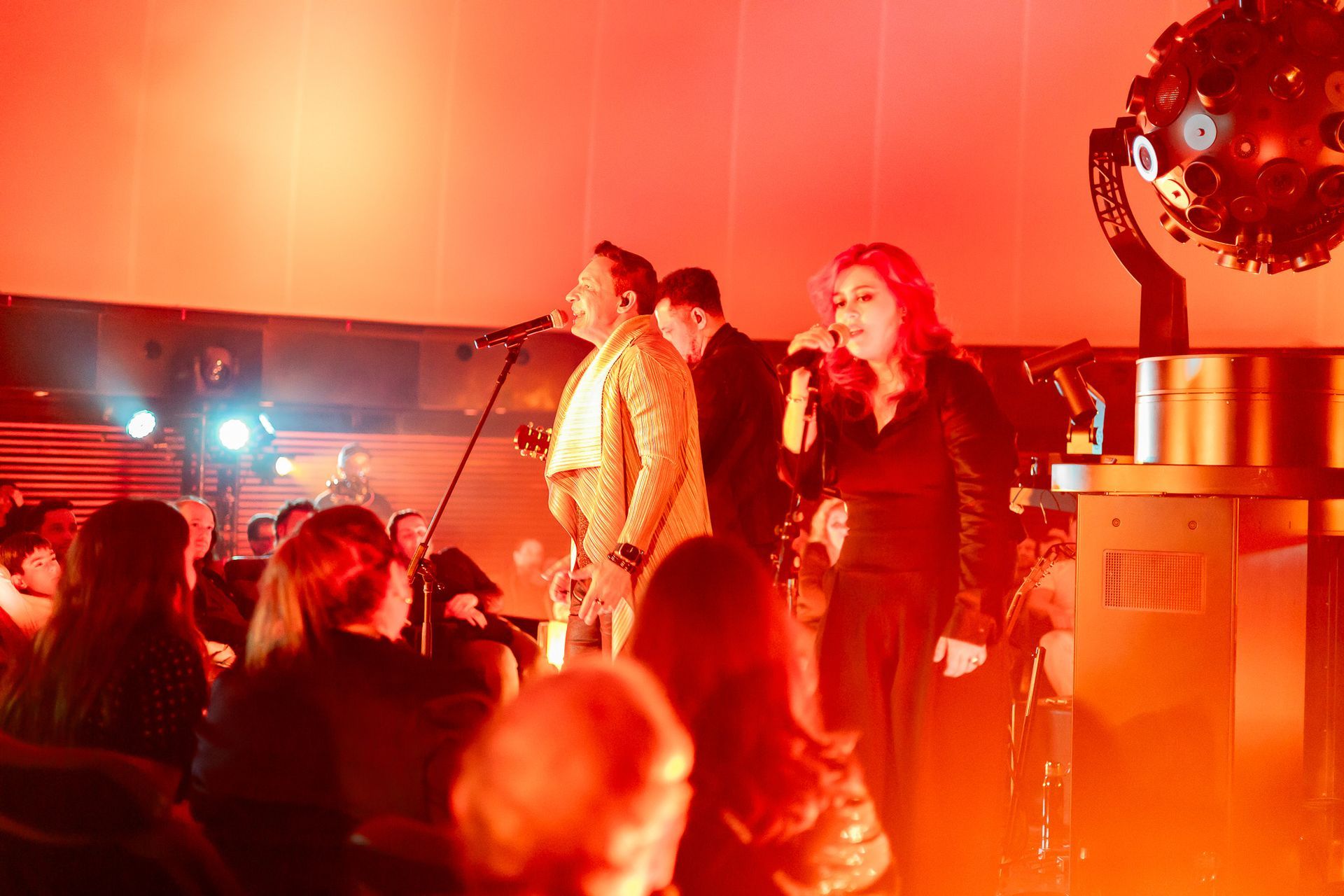Uma banda se apresenta em um palco iluminado  dentro de um planetário, com um grande projetor de estrelas visível