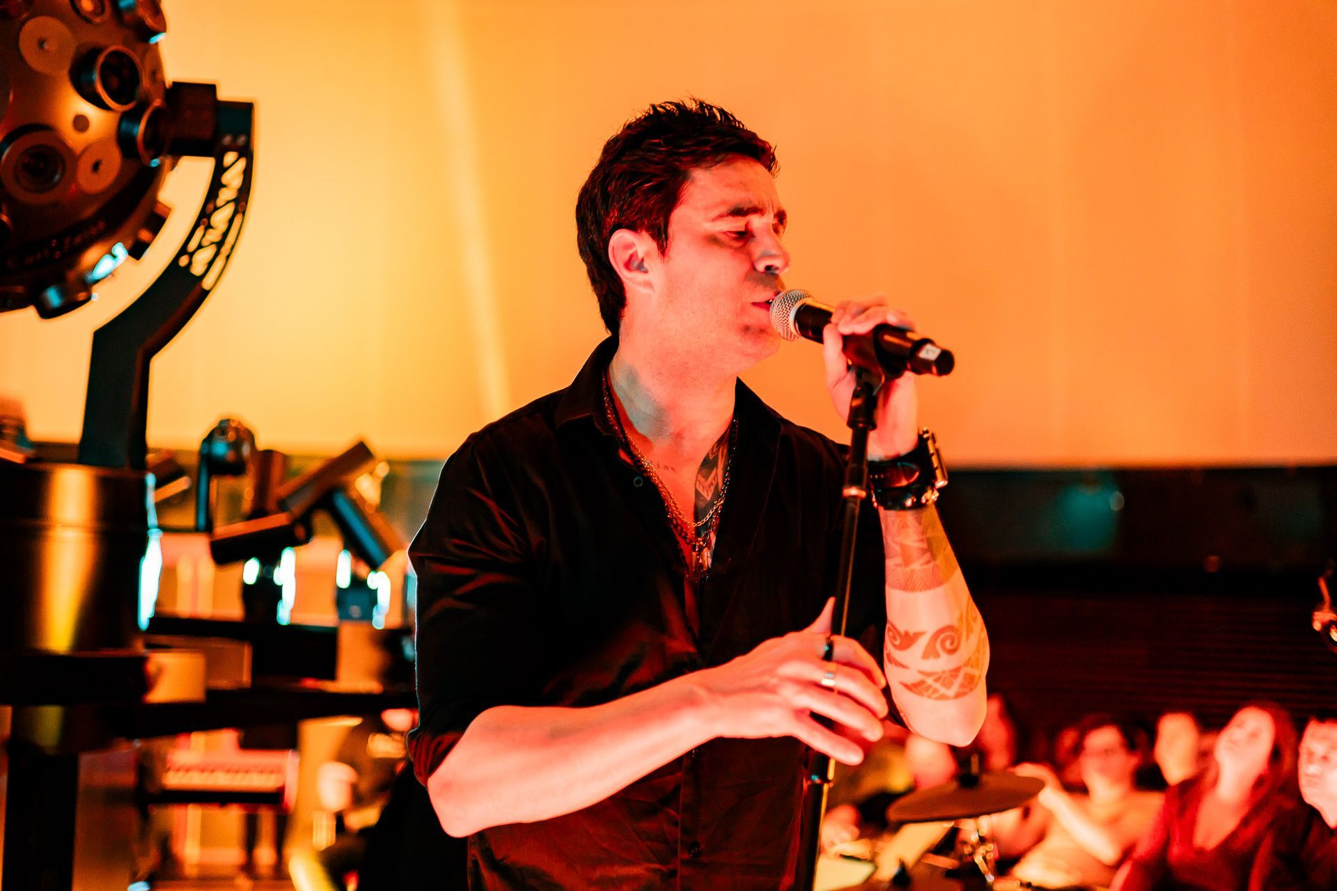 Um artista vestindo uma camisa preta canta em um microfone no palco, com um instrumento iluminado ao fundo e uma plateia.