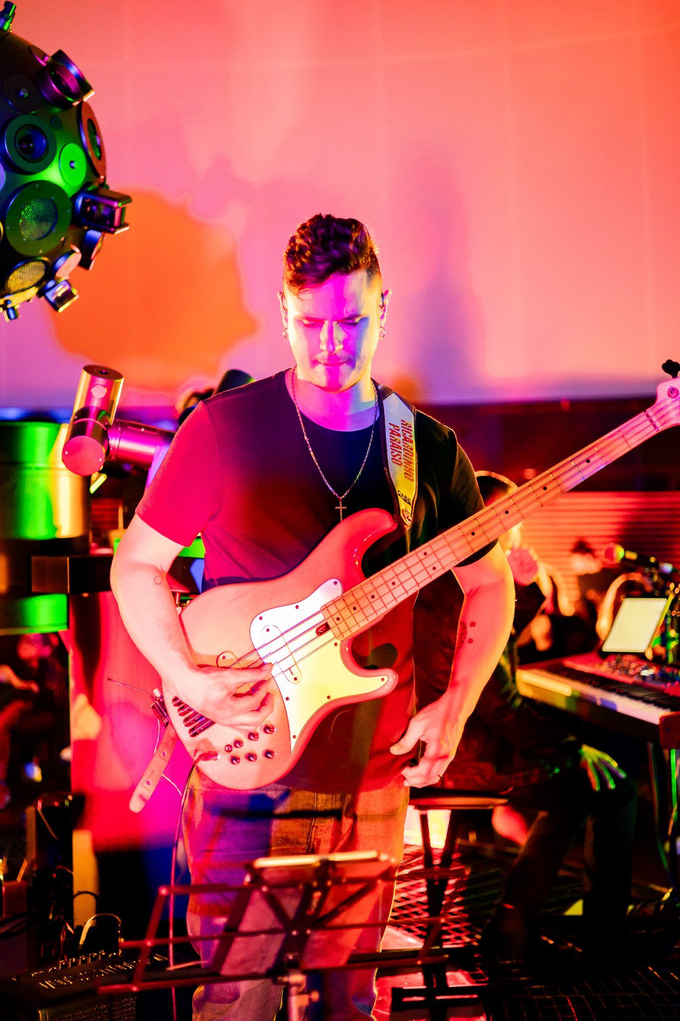 Uma pessoa tocando baixo em um palco pouco iluminado, com luzes de palco vermelhas e verdes vibrantes.