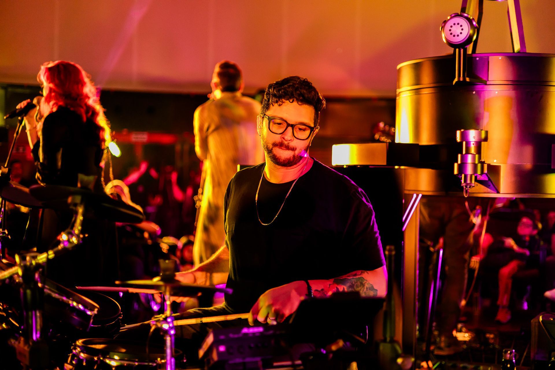 Um baterista de óculos se apresenta em um show sob uma iluminação quente e avermelhada, com outros membros da banda ao fundo.