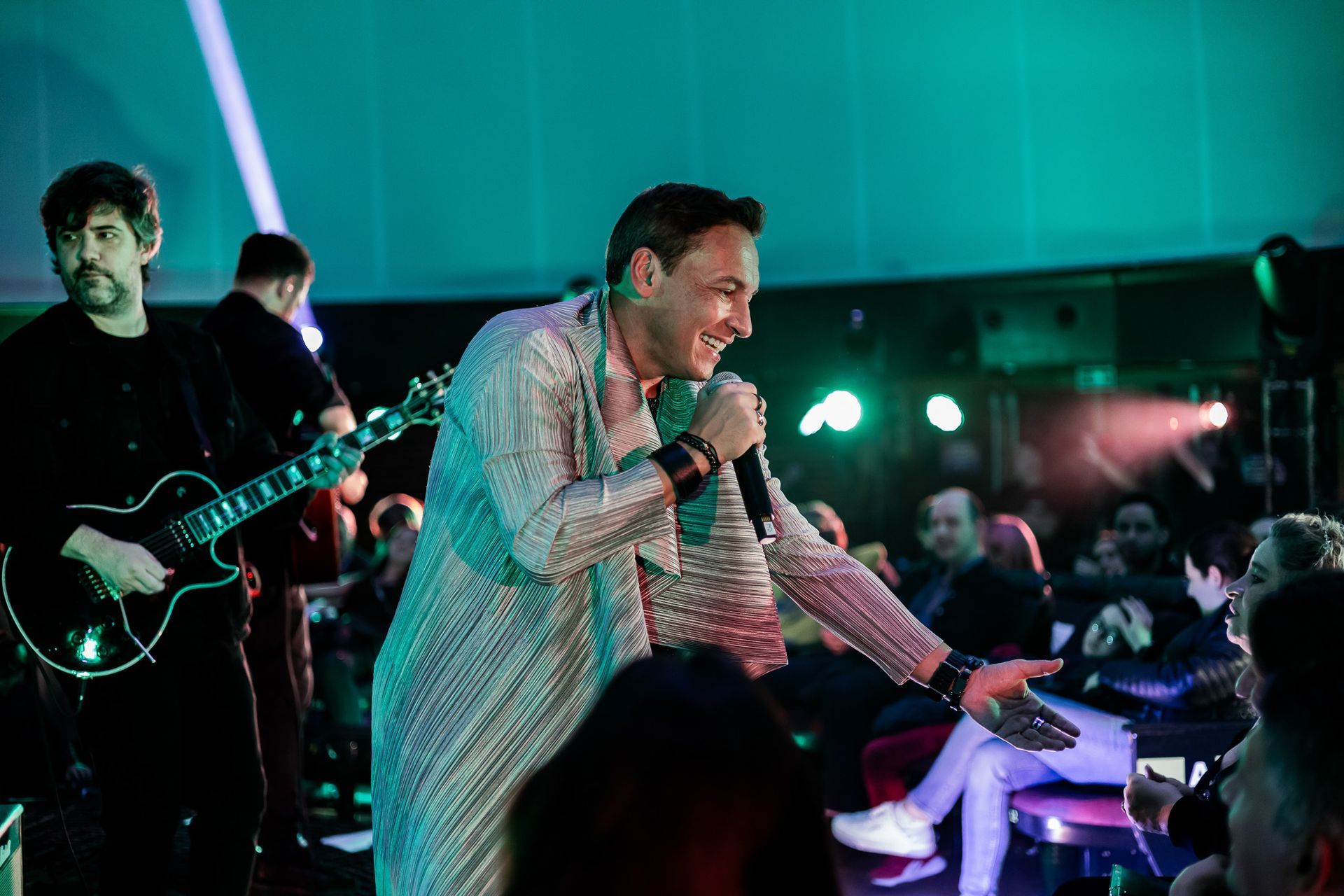 Um cantor vestido com um robe brilhante se apresenta no palco com um guitarrista, estendendo a mão em direção à plateia.