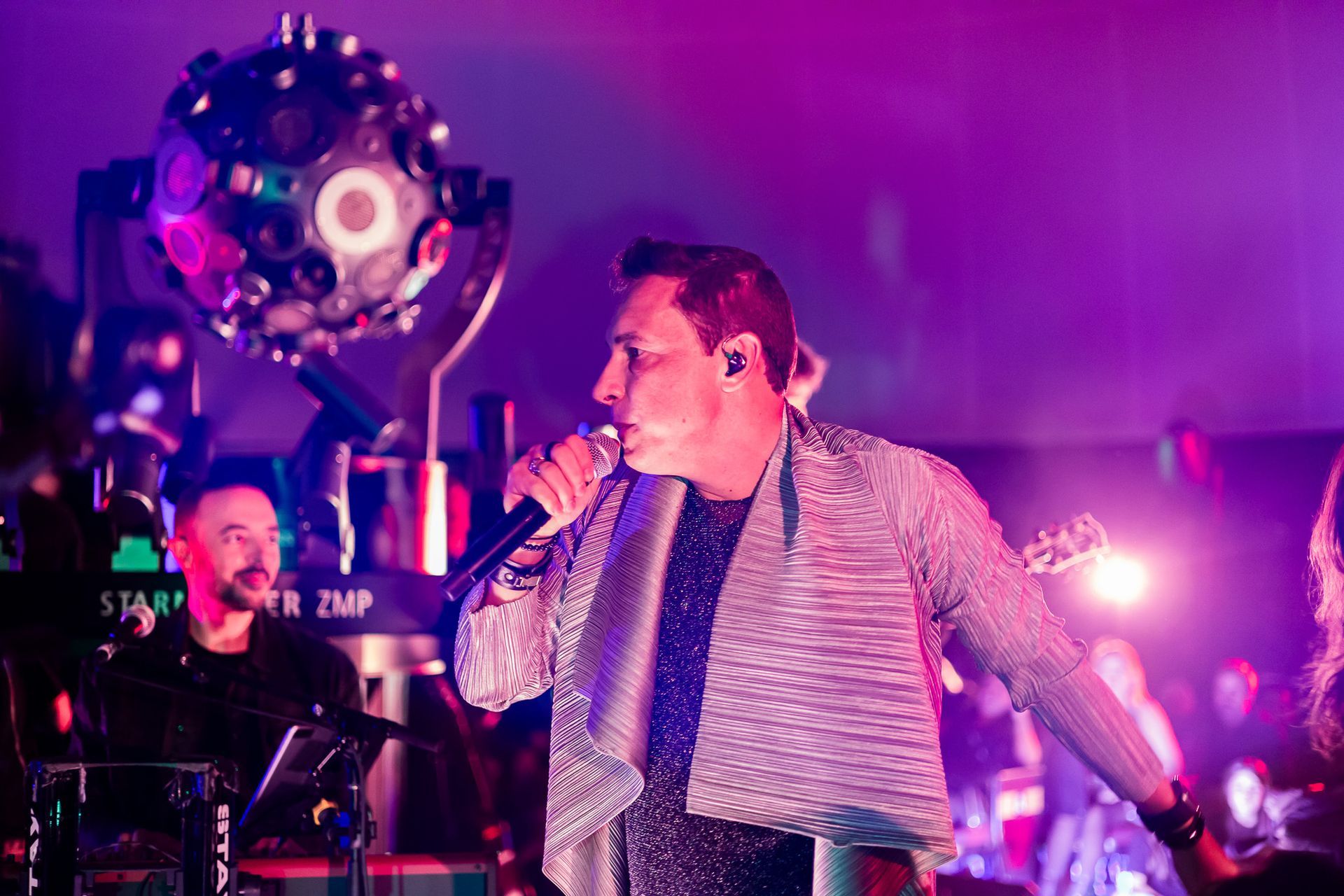 Um vocalista cantando em um microfone no palco, iluminada por luzes rosa e com um grande refletor esférico ao fundo.