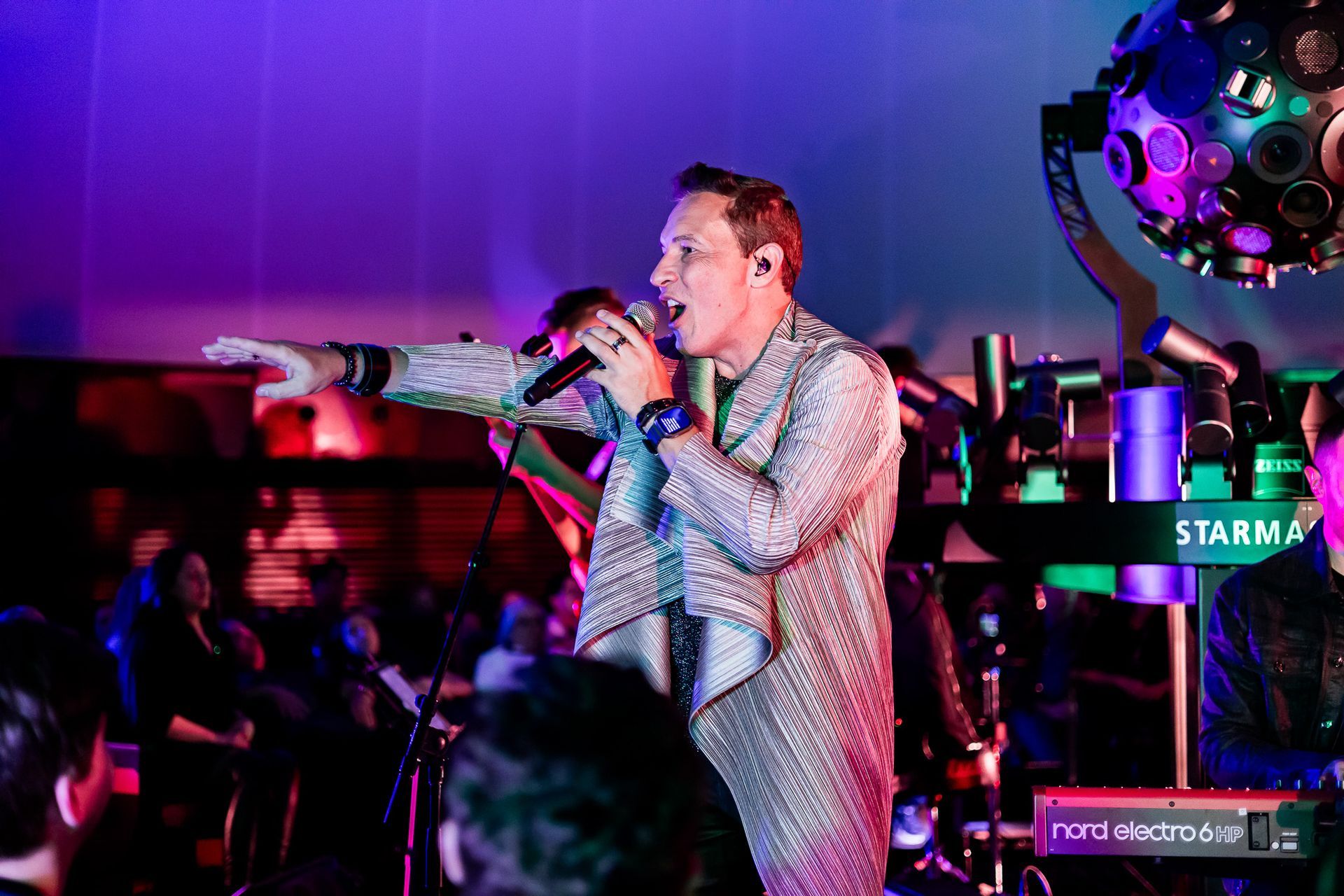 Um artista com uma jaqueta brilhante gesticula à plateia sob luzes de palco roxas vibrantes.