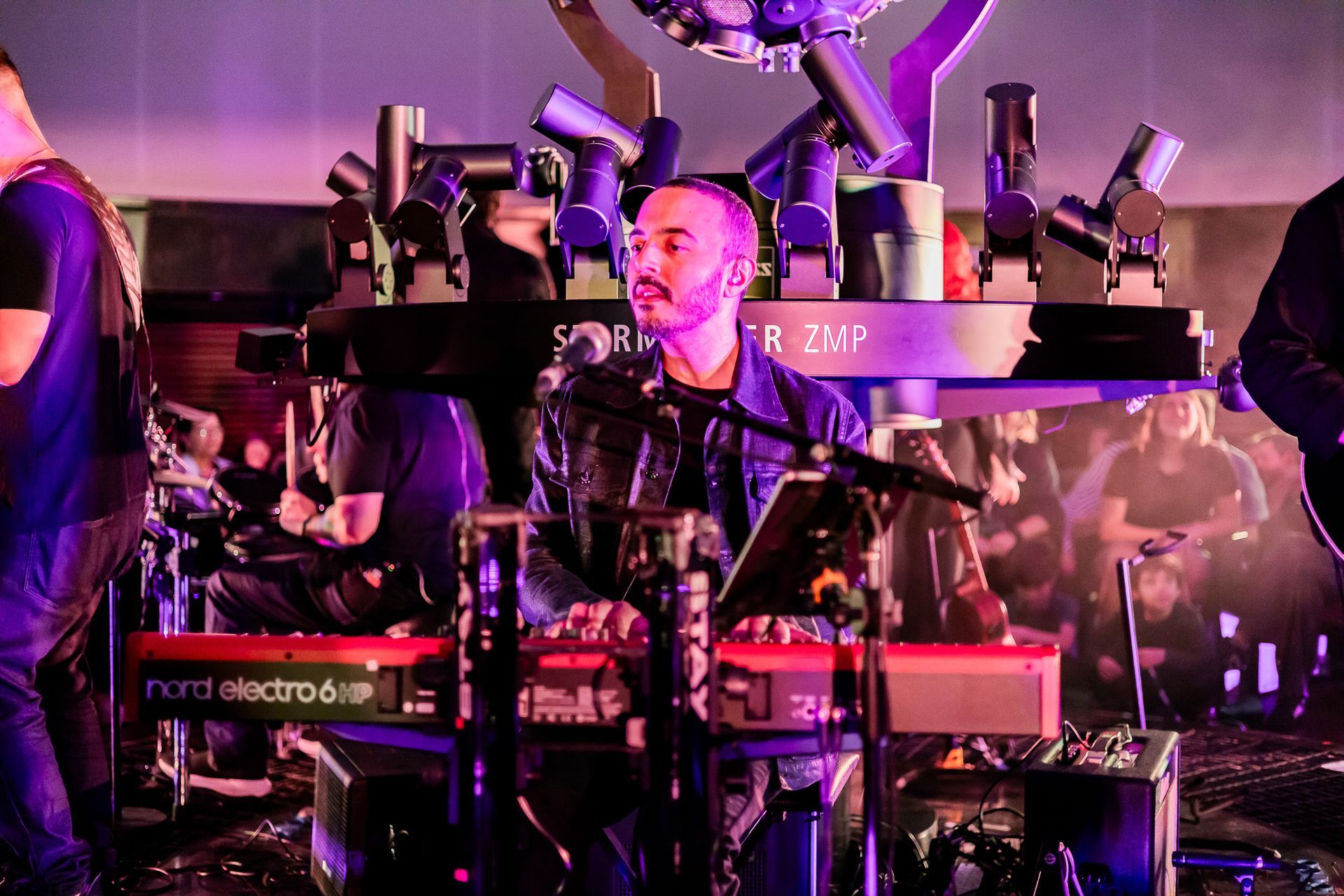 Um tecladista de jaqueta escura toca um teclado em um palco com iluminação em tons de roxo, iluminado por luzes de palco.