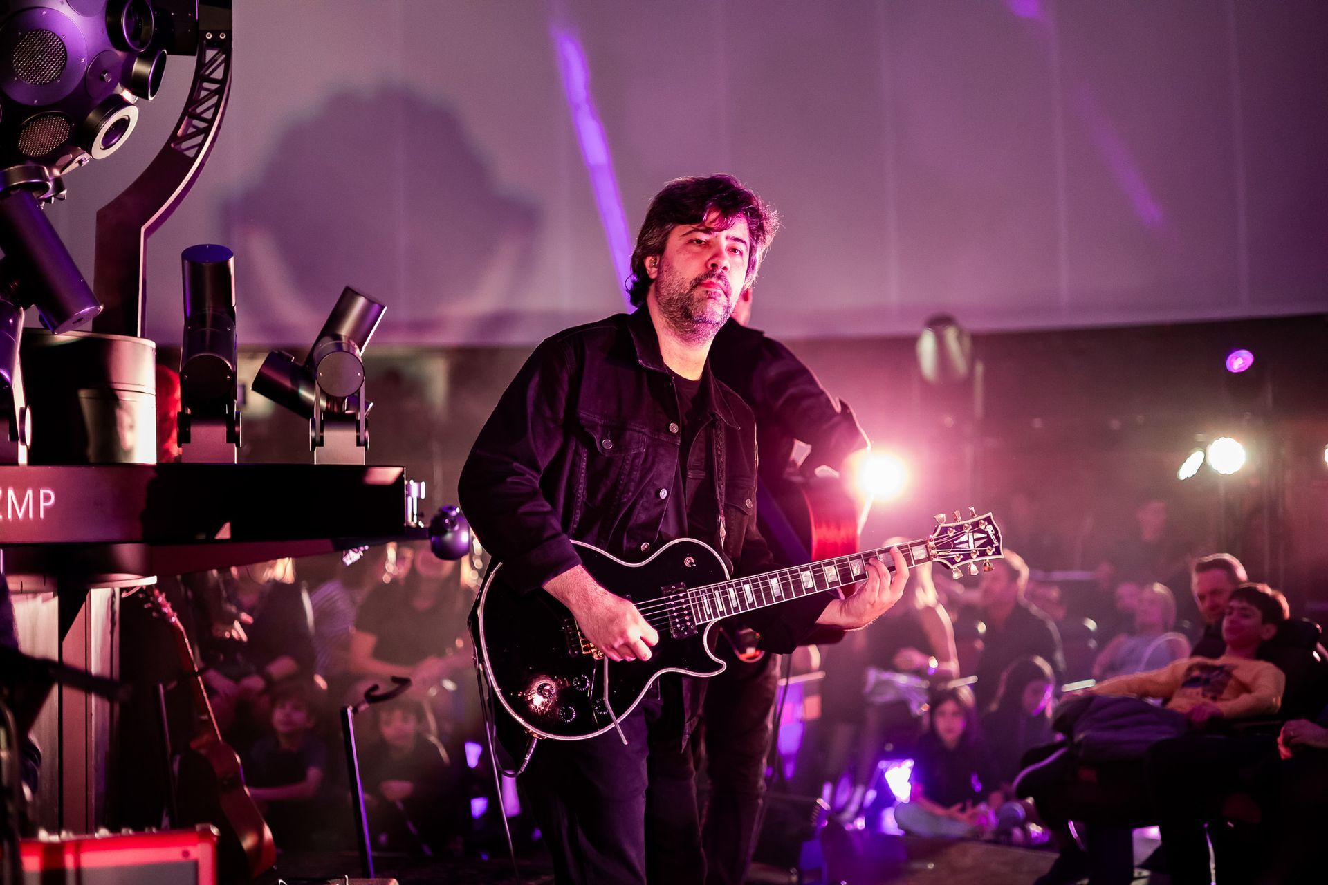 Um músico toca uma guitarra elétrica preta em um palco iluminado por luzes de palco rosa e roxas brilhantes.