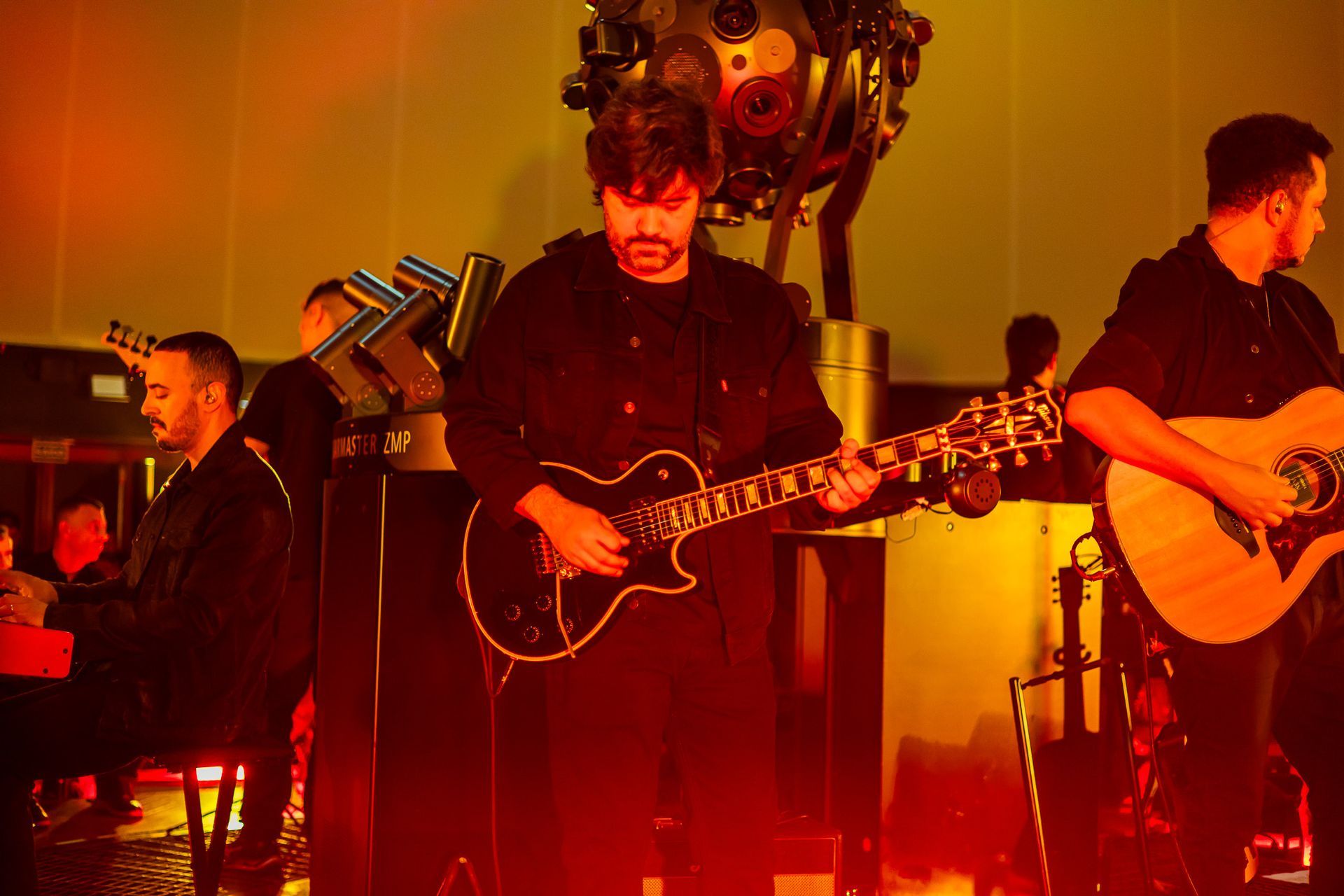 Uma banda se apresenta em um palco com iluminação aconchegante em tons de laranja. Os músicos tocam guitarra e teclado.