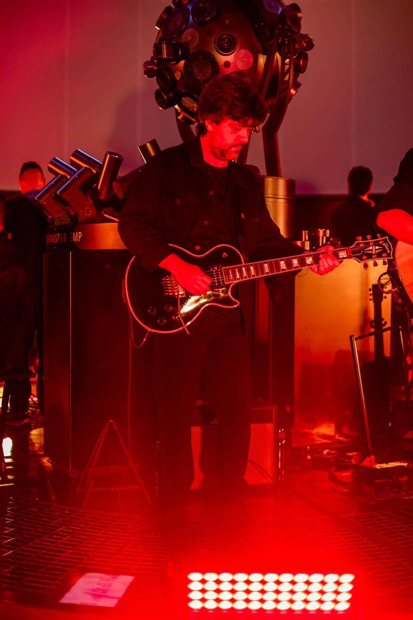 Um guitarrista se apresenta no palco sob uma forte iluminação vermelha, cercado por equipamentos musicais e sombras.