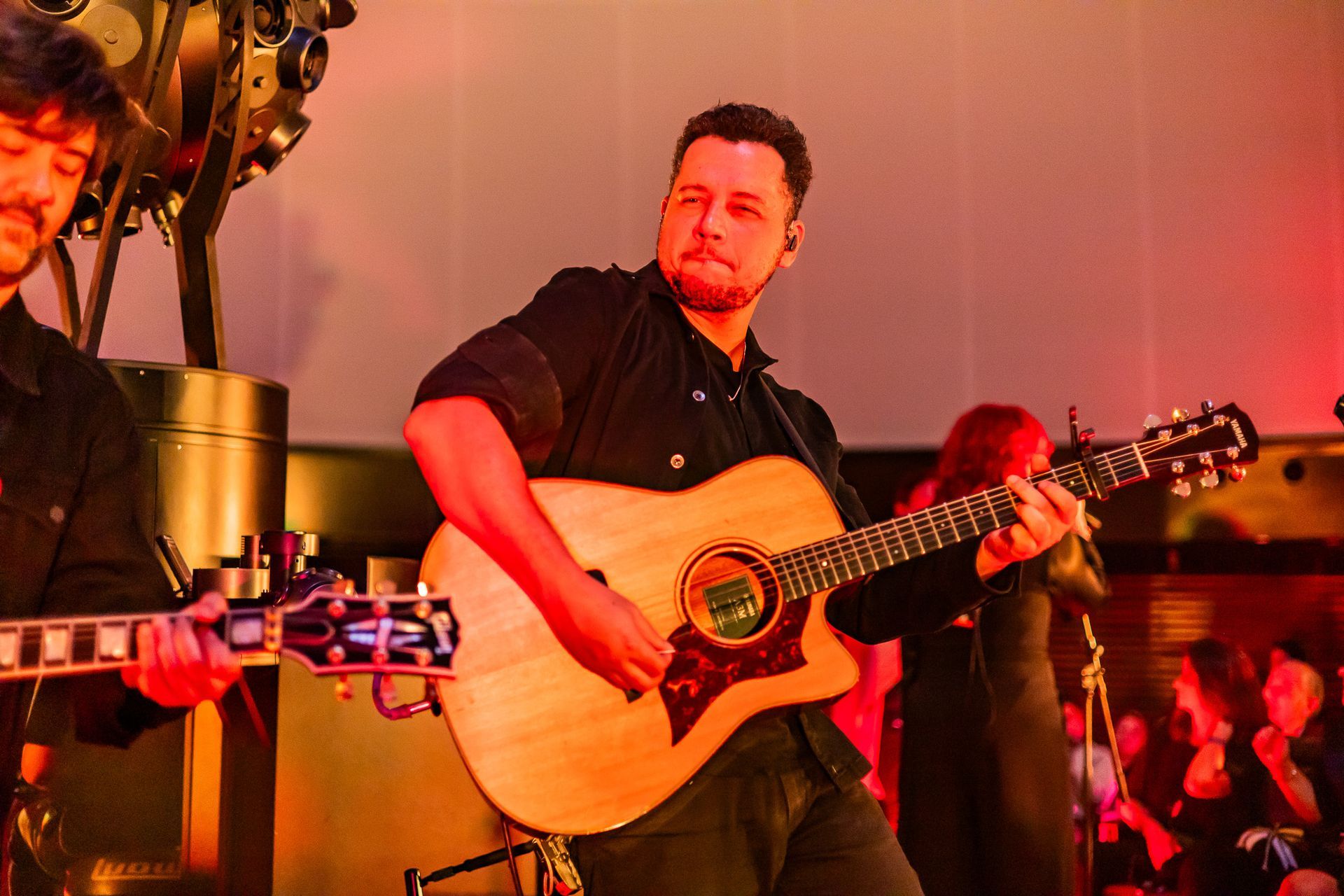 Um músico tocando violão enquanto se apresenta no palco sob uma iluminação de concerto quente, em tons de vermelho.