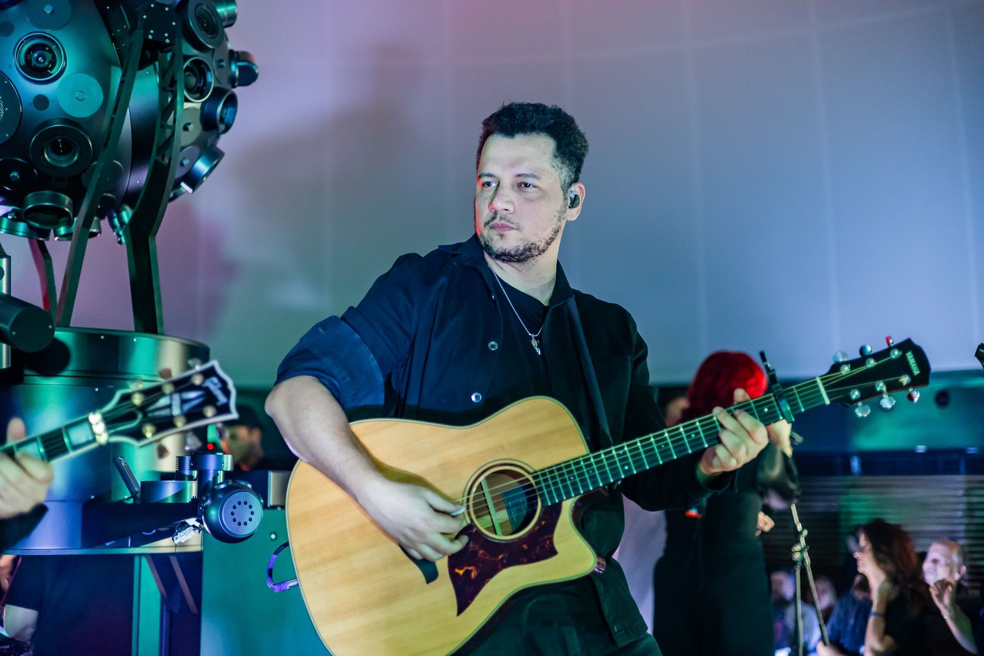 Um artista tocando violão em um palco, com um equipamento de câmera com múltiplas lentes visível em primeiro plano.