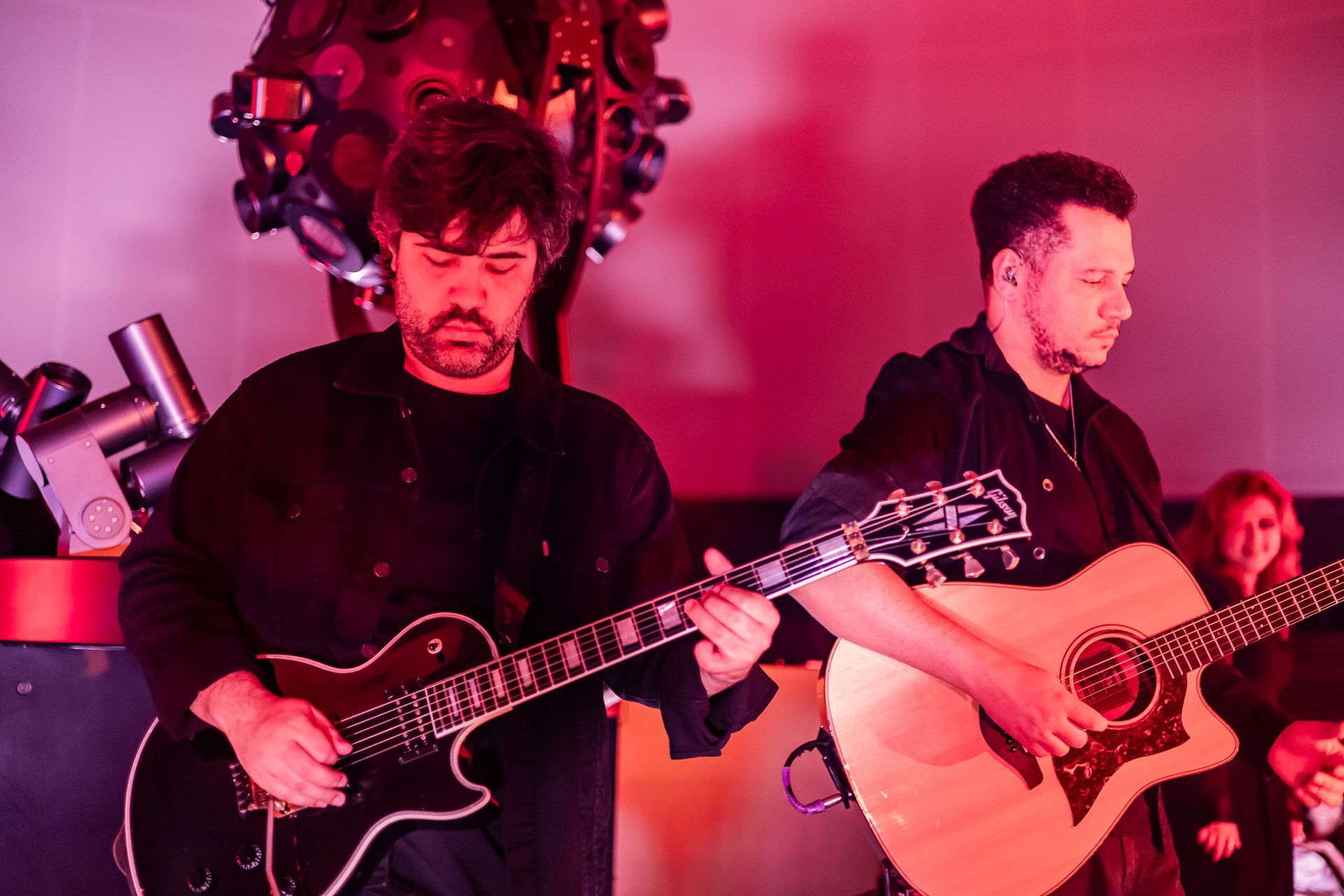 Dois músicos se apresentam sob luzes vermelhas, um tocando guitarra elétrica e o outro tocando violão.