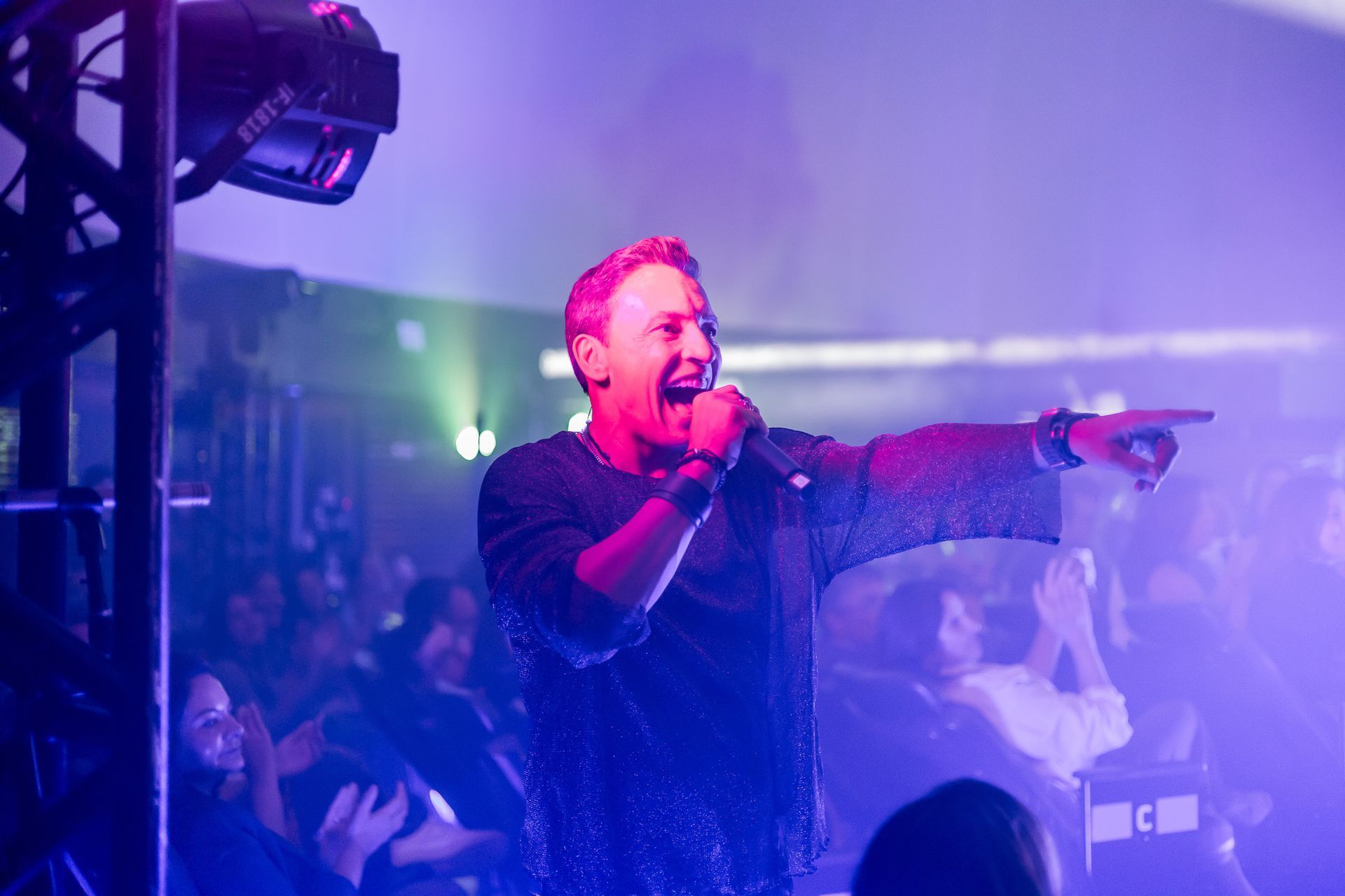 Um artista canta em um microfone no palco, apontando para a plateia sob uma vibrante iluminação roxa de concerto.
