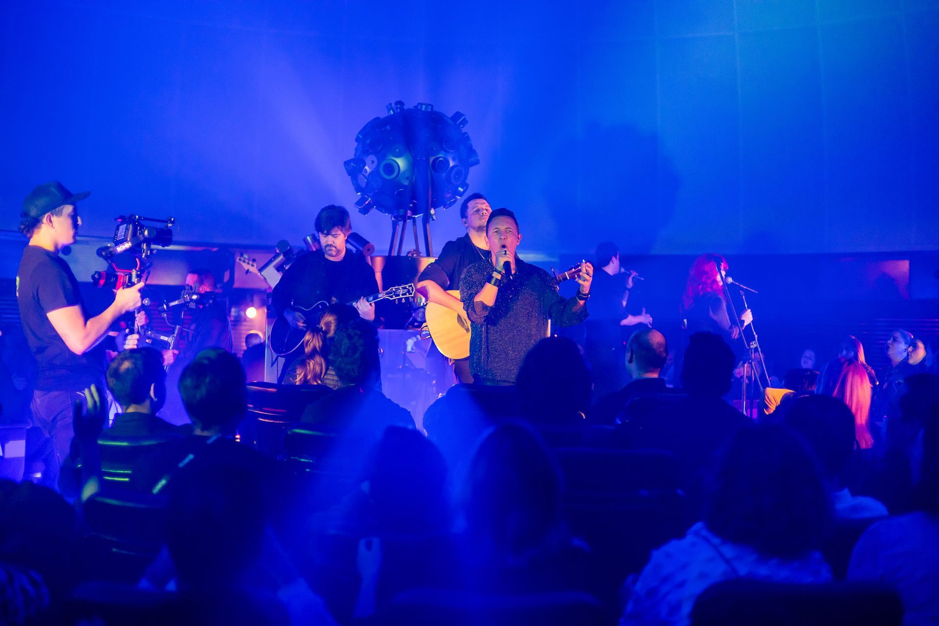 Uma banda se apresenta no palco sob holofotes azuis, com um operador de câmera em primeiro plano e o público no escuro.