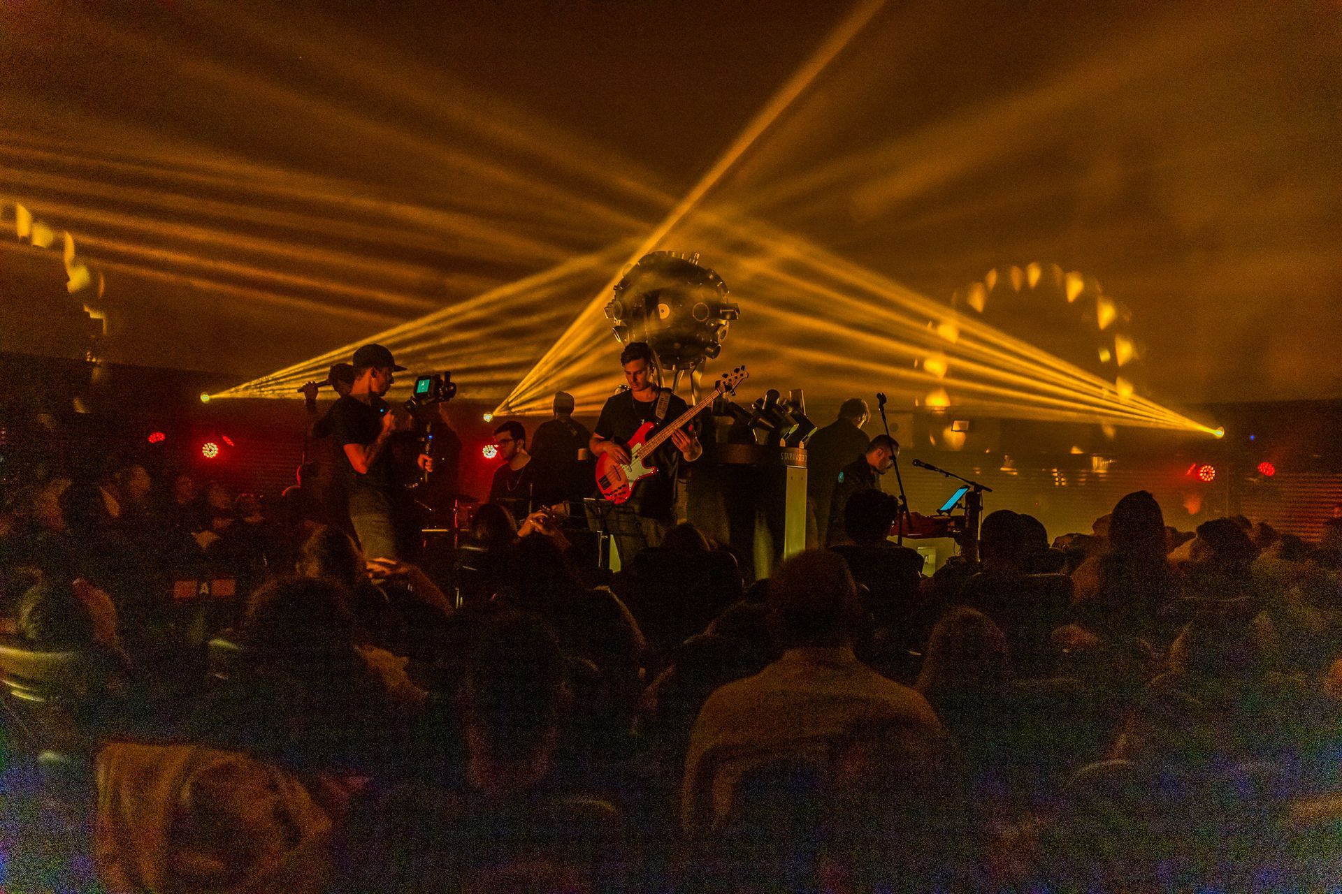 Uma banda se apresenta no palco sob holofotes dourados e uma bola de discoteca, sua silhueta projetada diante de uma plateia