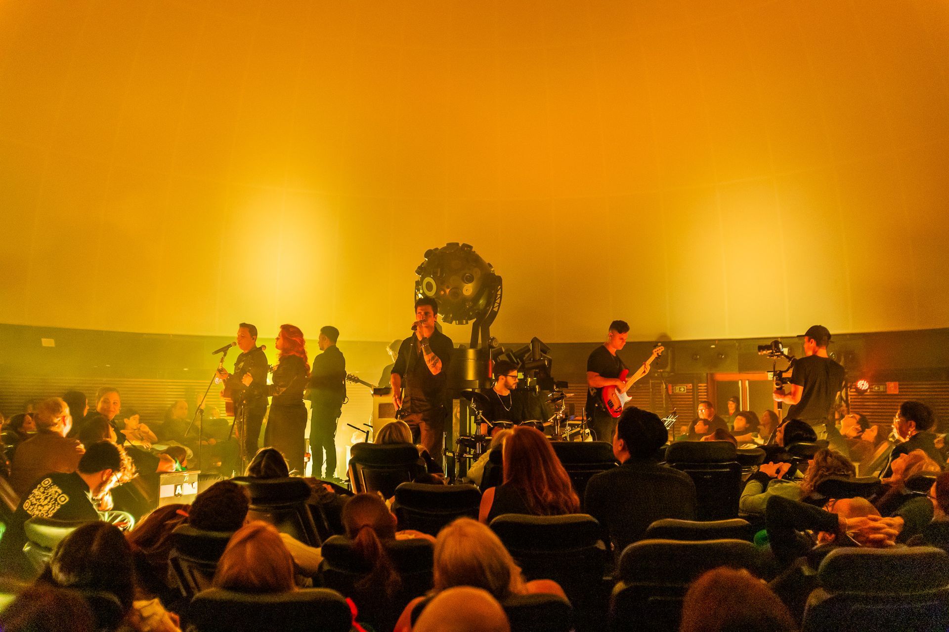 Uma banda se apresenta em um palco dentro de um teatro, iluminado por uma luz dourada, vista da área reservada à plateia.
