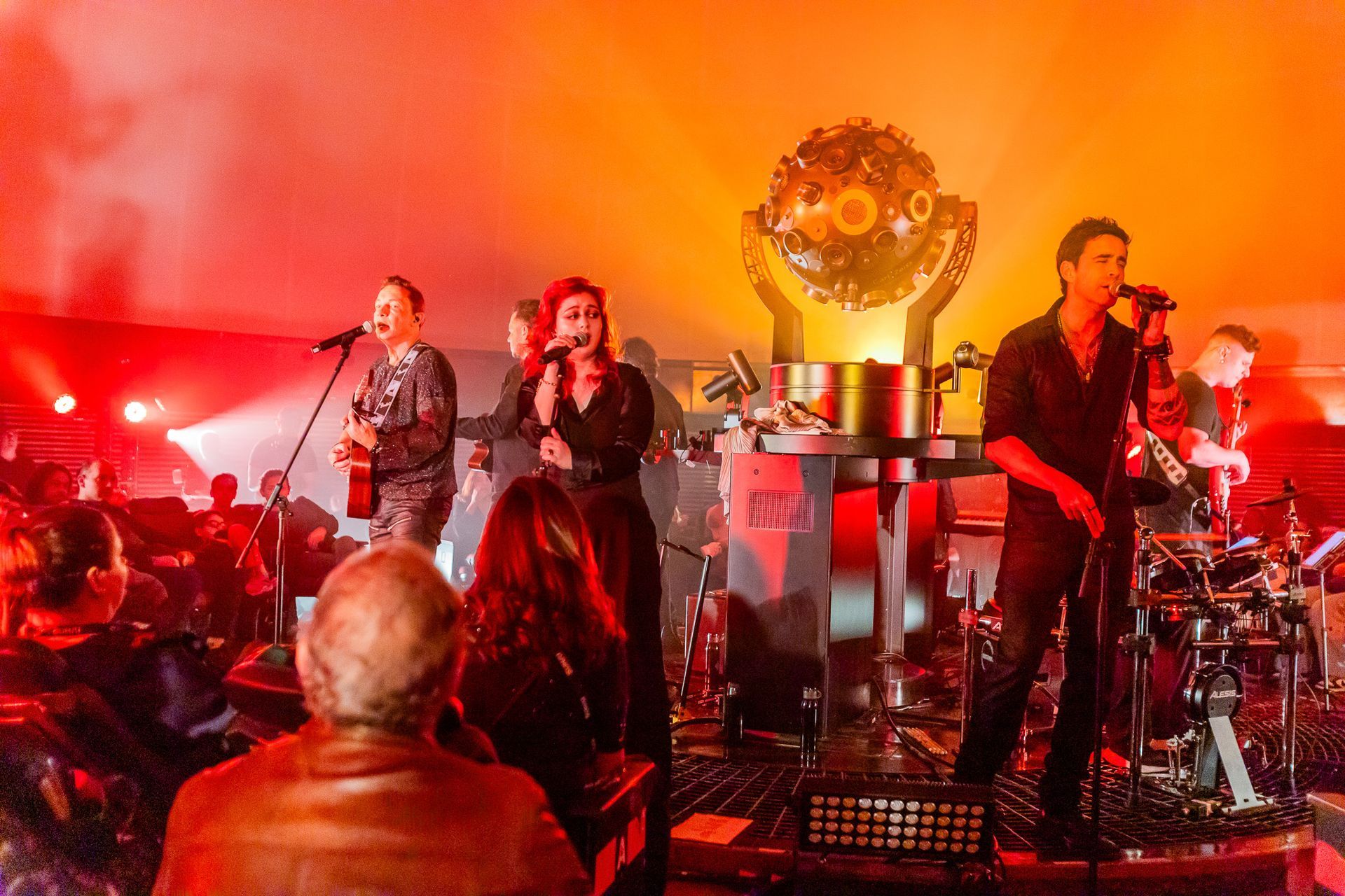 Uma banda se apresenta em um palco, com uma escultura de percussão central e uma plateia sentada à frente.