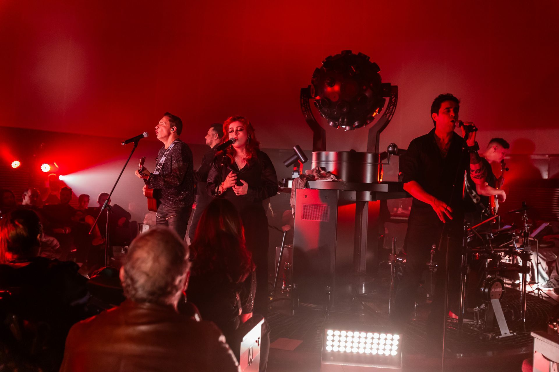 Uma banda se apresenta em um palco iluminado com luz vermelha, composta por vocalistas, um guitarrista e um baterista.