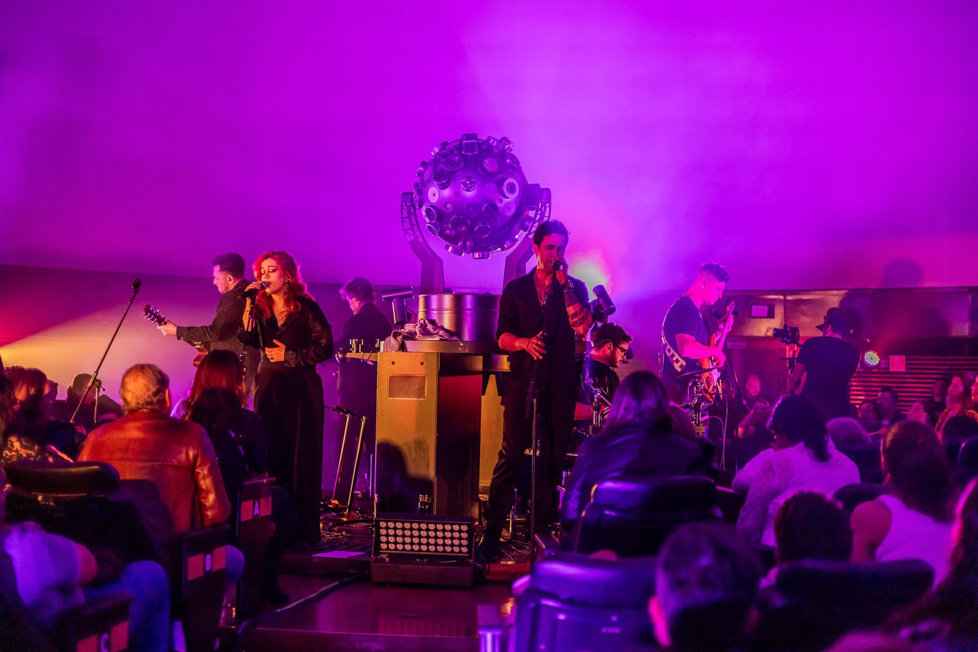 Uma banda se apresenta no palco sob uma iluminação roxa intensa, diante de uma plateia sentada em um local fechado e escuro.