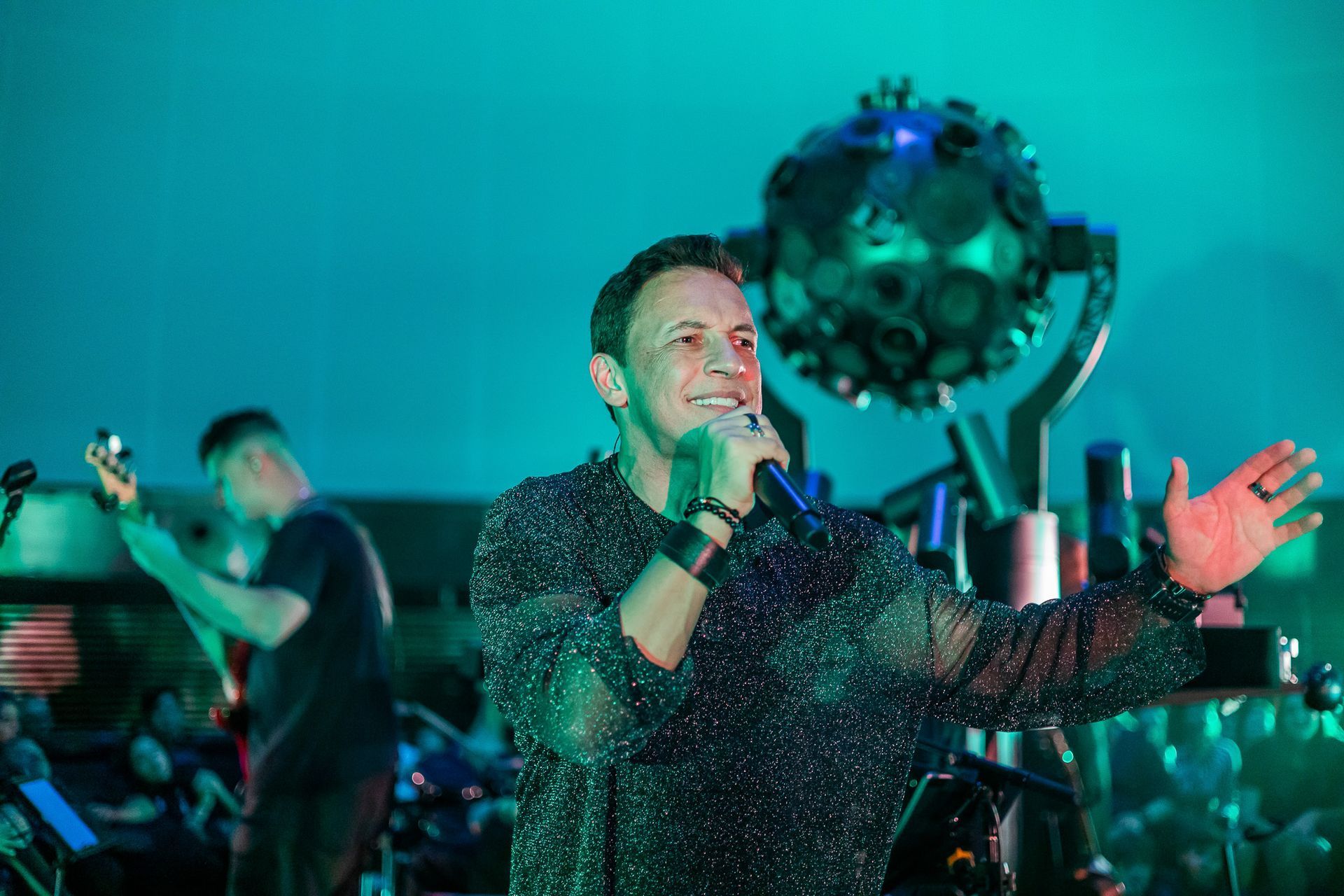 Um artista cantando em um microfone no palco, iluminada, com uma grande esfera estilo bola de discoteca atrás dela.
