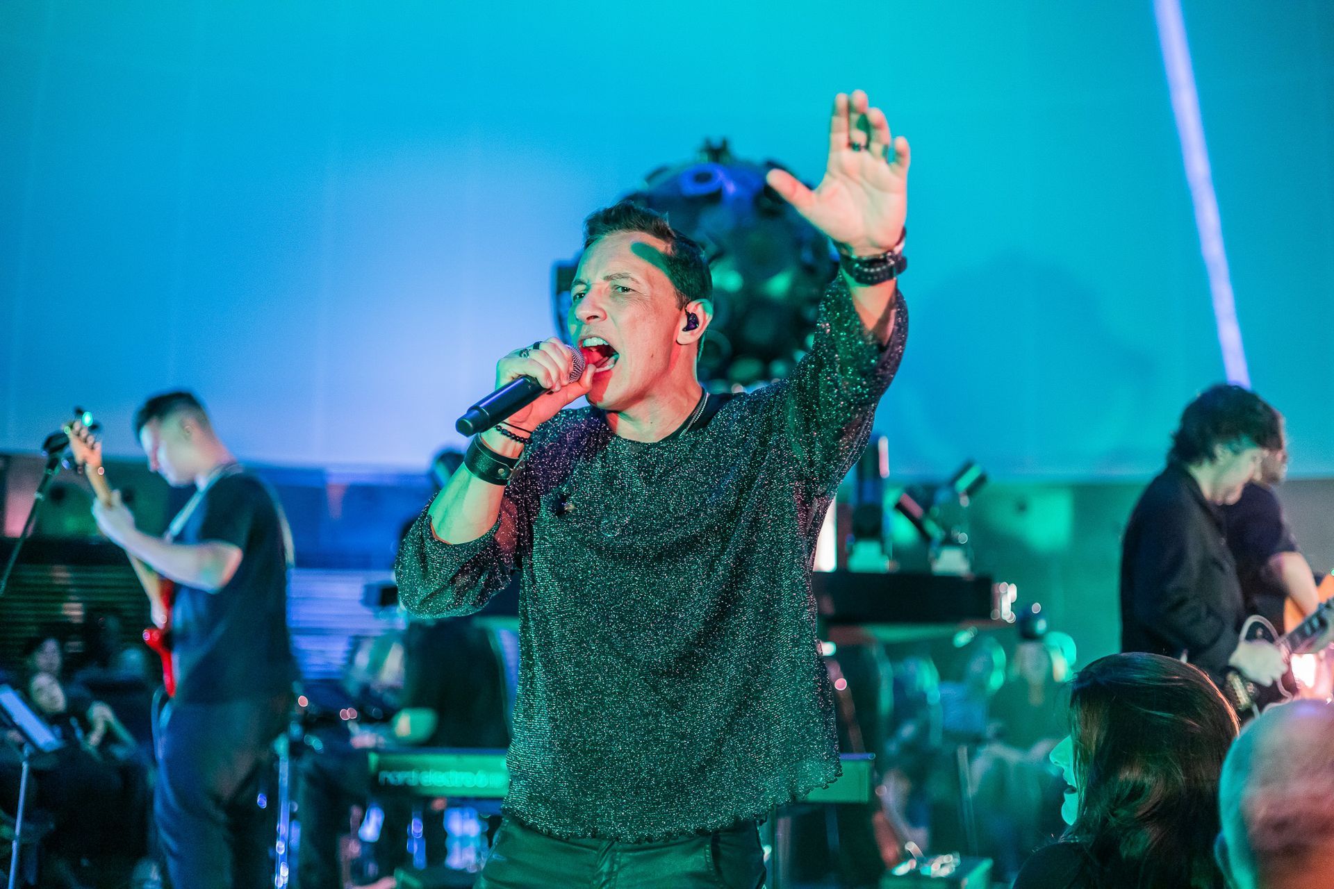 Um cantor se apresenta no palco com uma banda sob iluminação azul, segurando um microfone e gesticulando com a mão levantada.