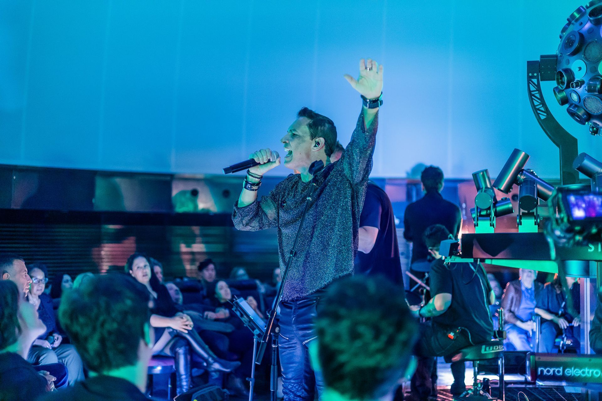 Um cantor com uma camisa estampada se apresenta no palco com a mão levantada, diante de uma plateia.