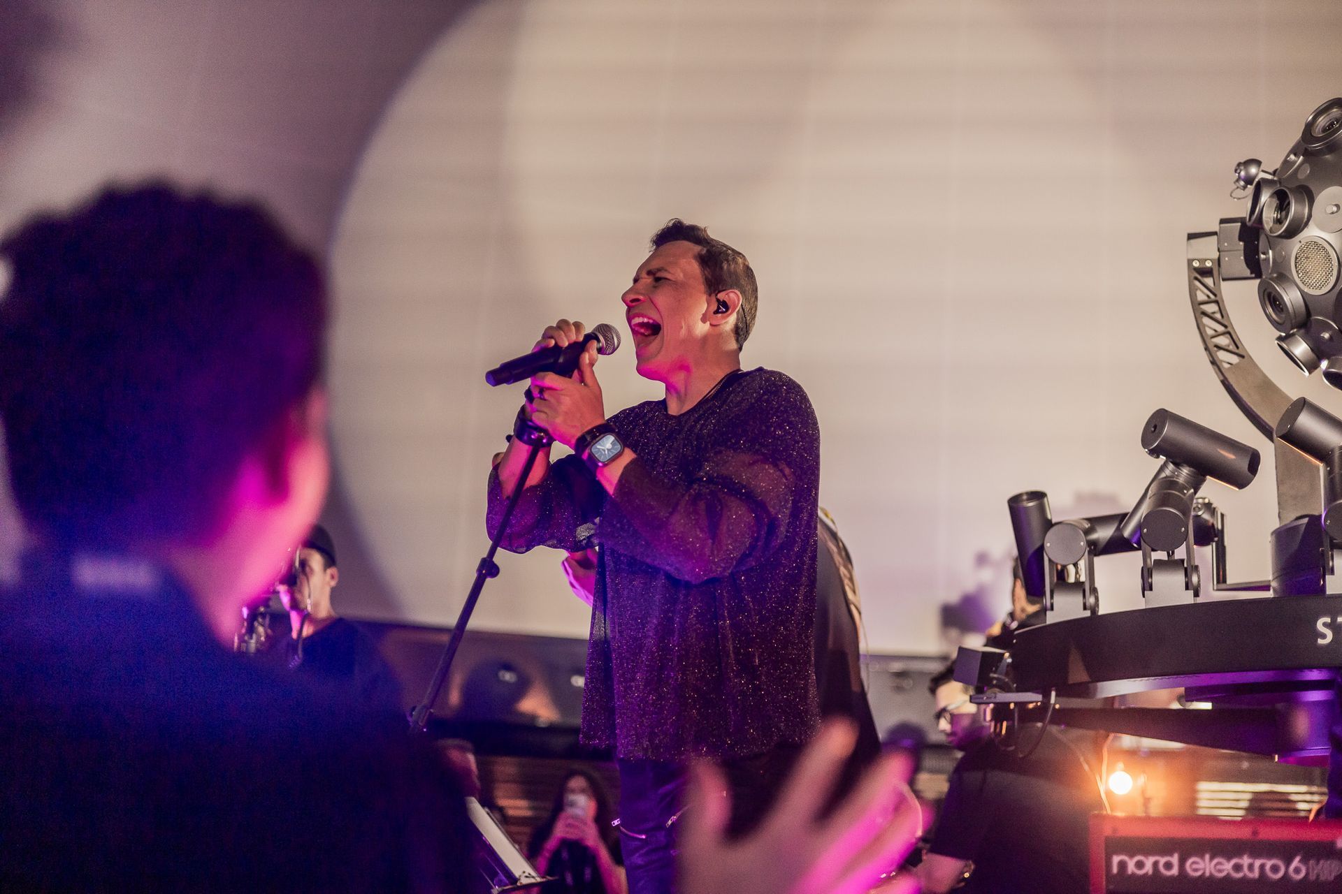 Um artista vestindo uma blusa escura e texturizada canta com paixão em um microfone no palco, iluminada por um holofote.
