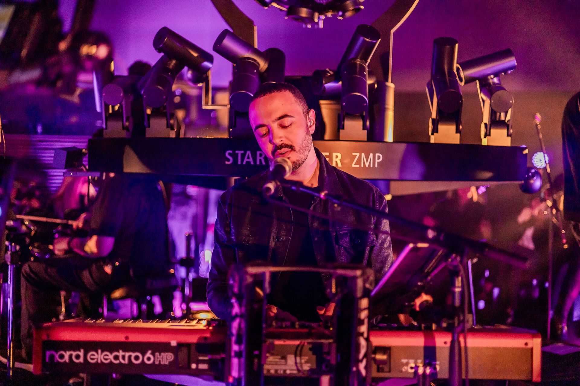 Um músico toca teclado sob luzes de palco roxas, cantando em um microfone em frente a uma estrutura de iluminação.