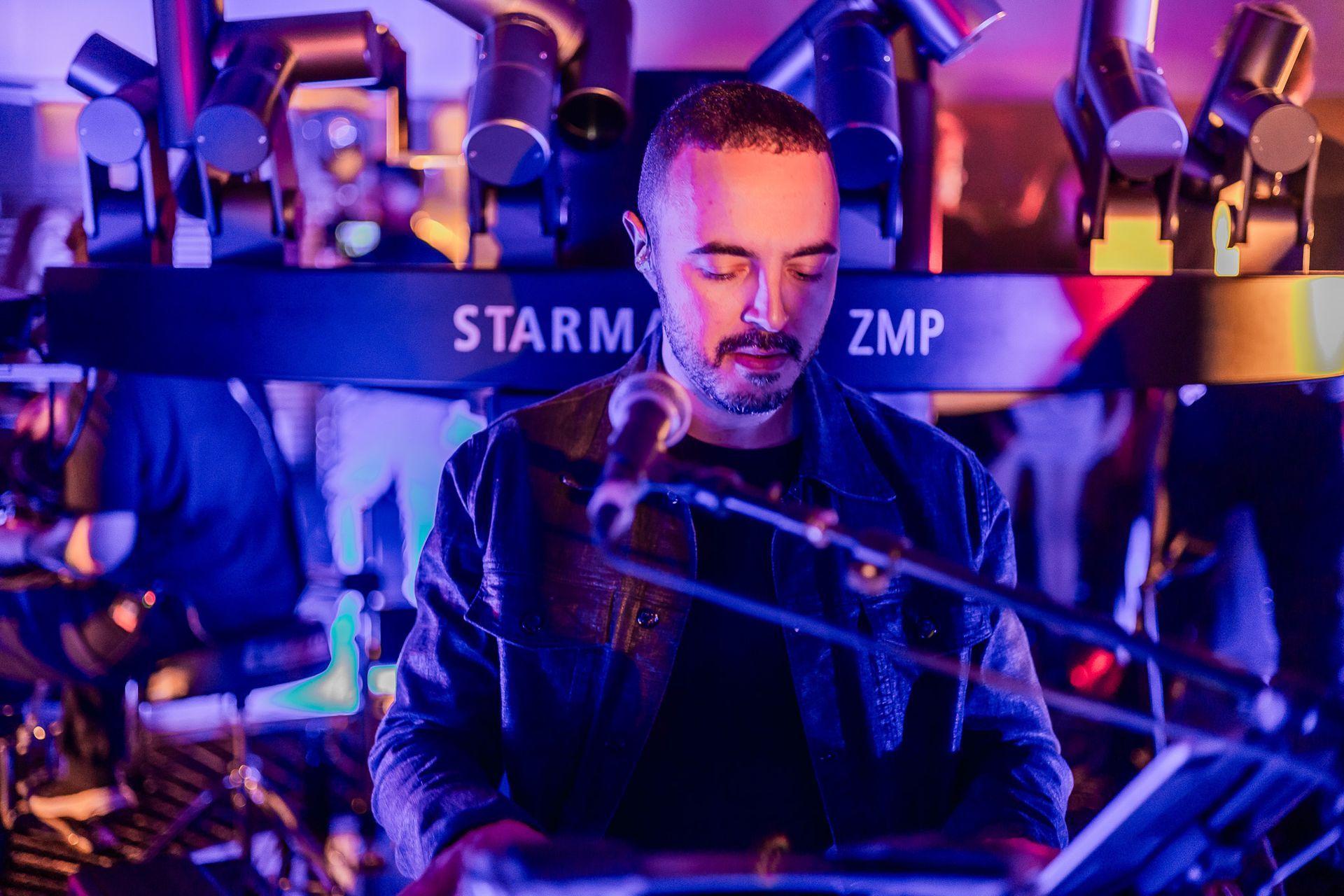 Um músico de jaqueta preta cantando ao microfone no palco, iluminado por luzes de concerto azuis e roxas.