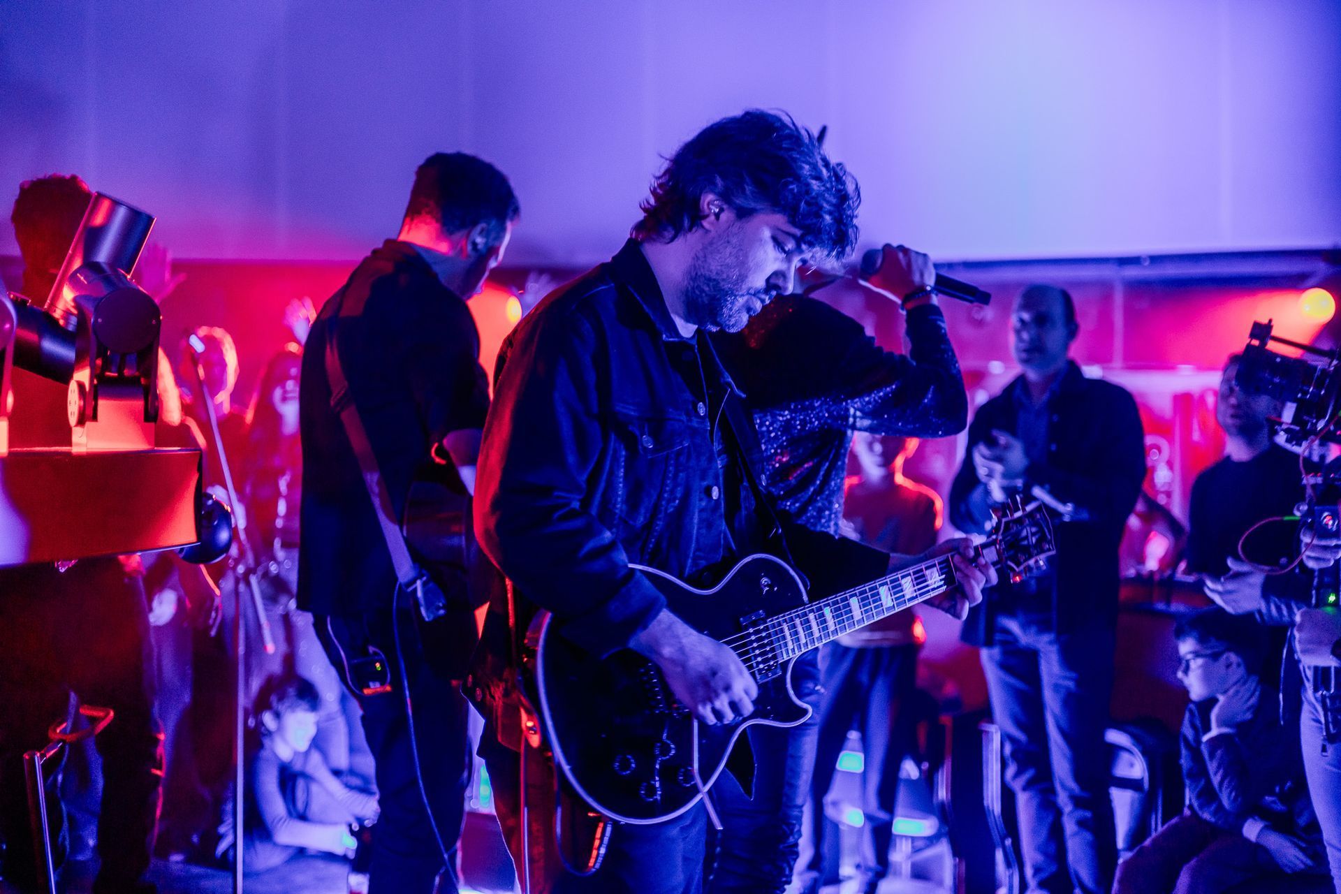 Um músico toca guitarra elétrica e canta em um microfone em um palco iluminado com luzes azuis e vermelhas.